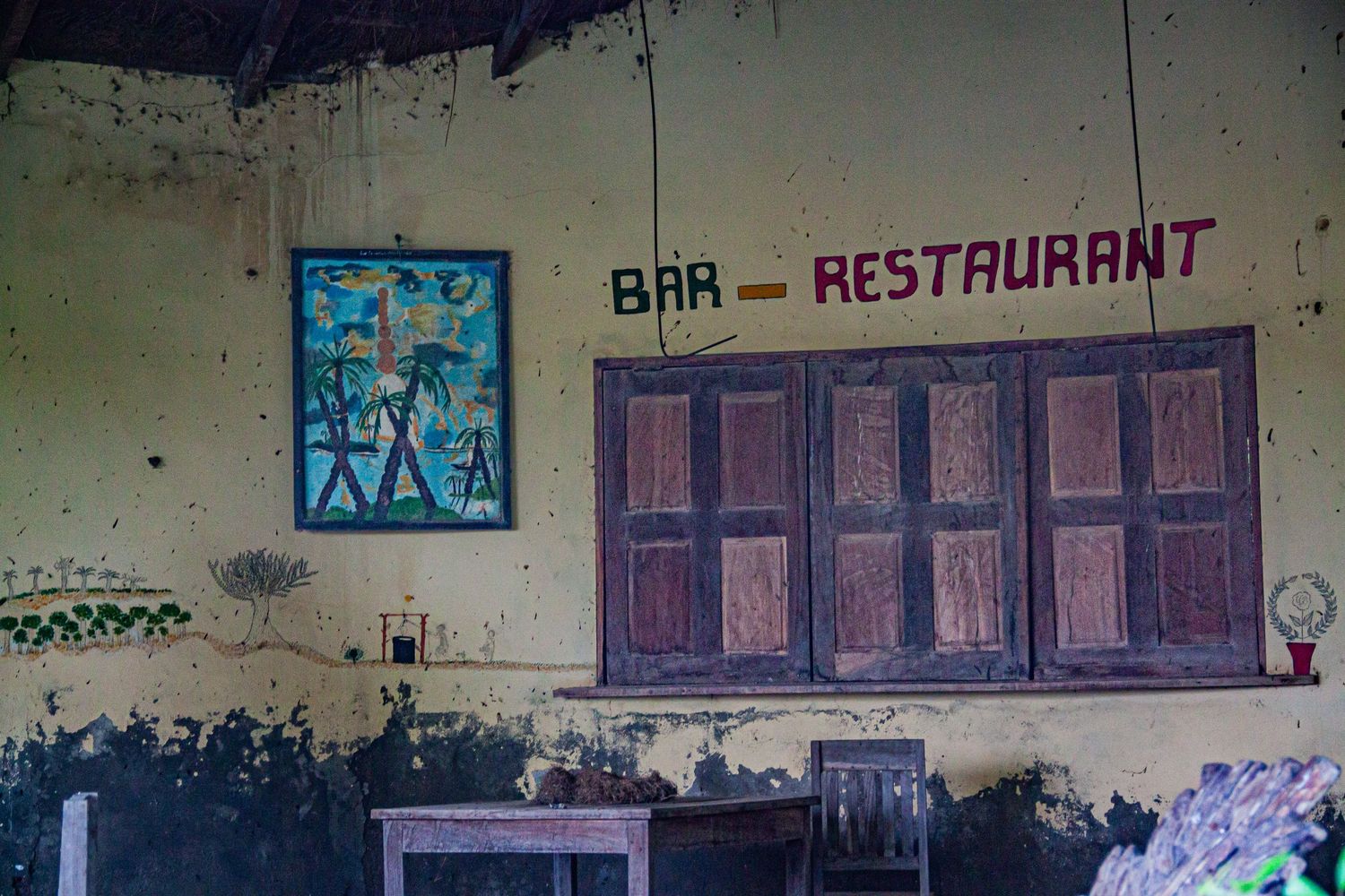 Rustic bar-restaurant exterior with yellow walls, wooden shutters, and colorful artwork in Africa.