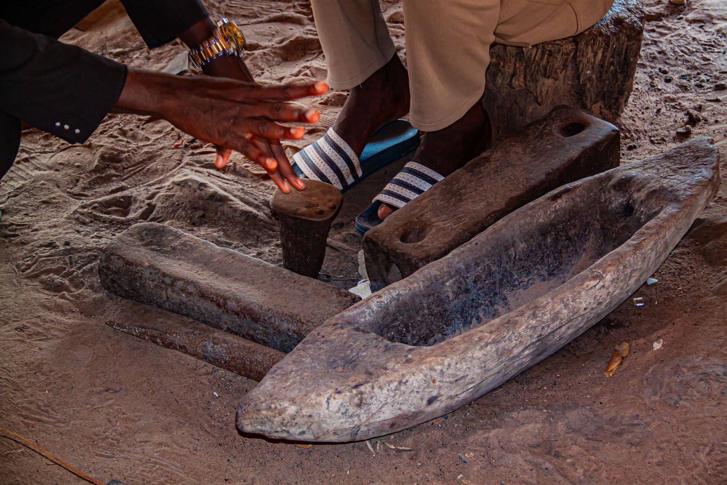 Hands examining ancient carved stone artifacts including a boat-shaped relic on sandy ground in Africa.