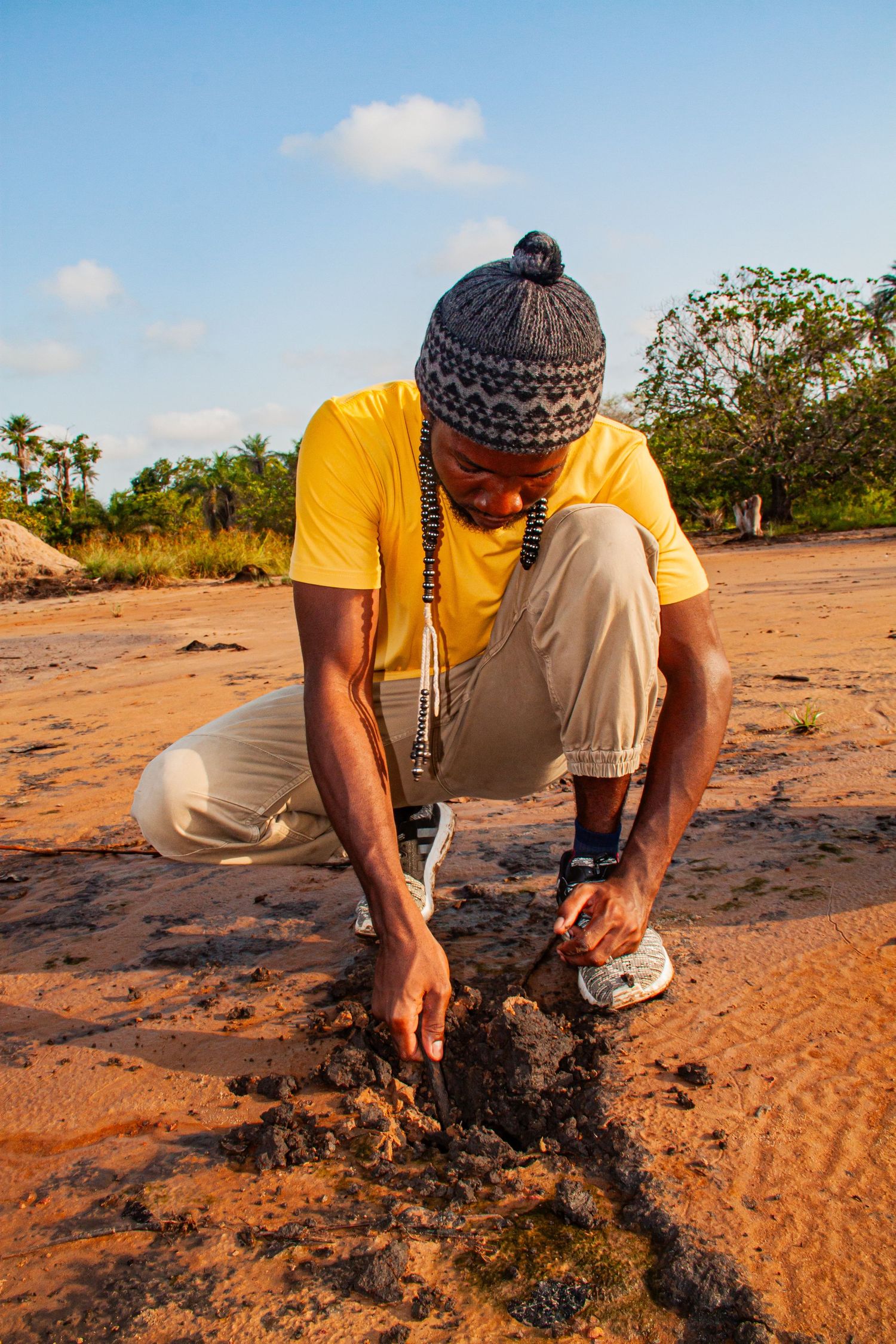 African man in yellow shirt and knit hat crouching on sandy ground examining soil.