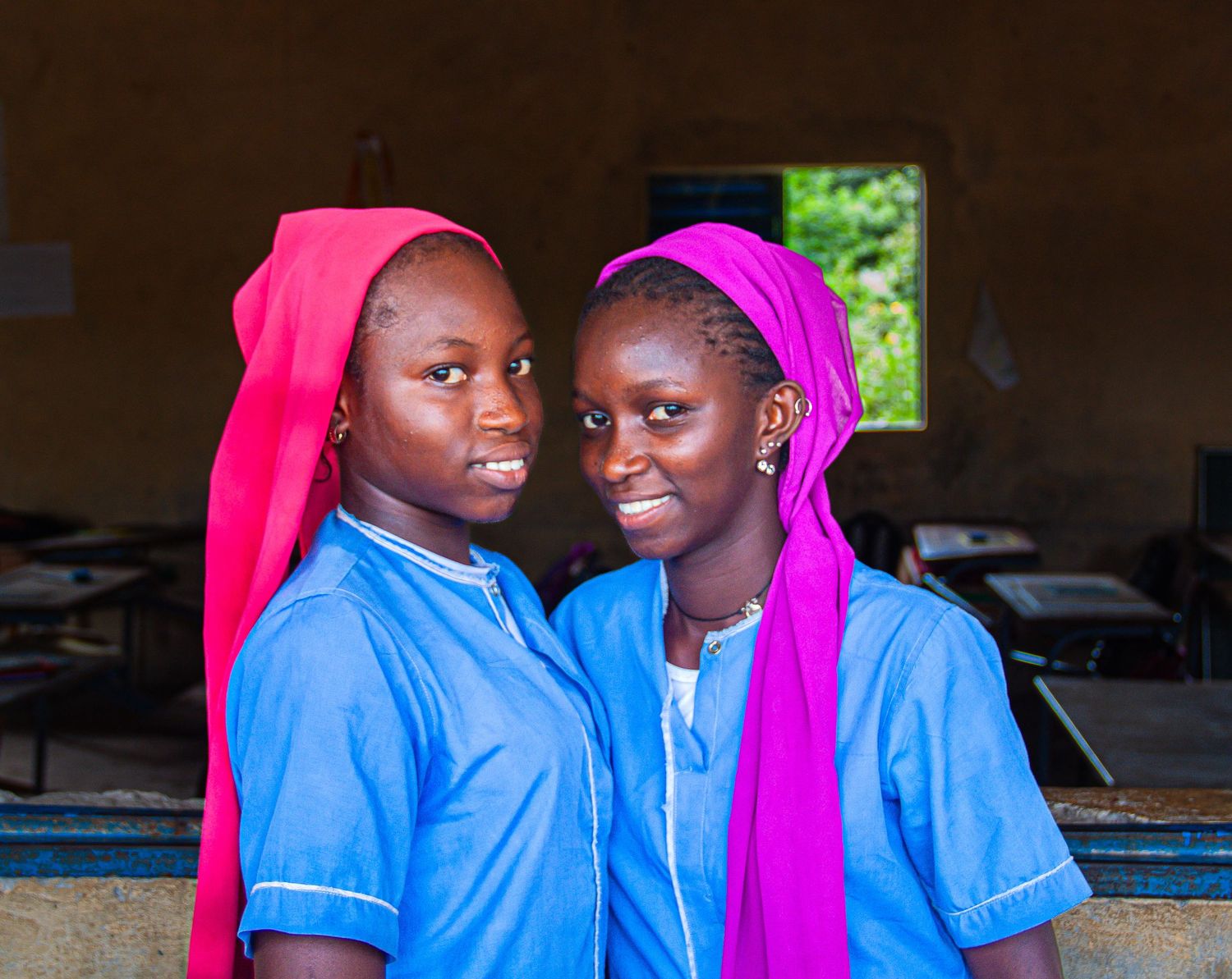 Two young African girls in blue uniforms and pink headscarves smiling in a classroom.
