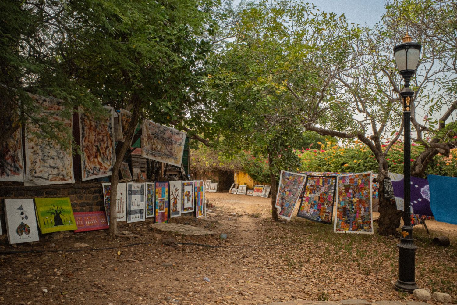 Colorful African paintings displayed outdoors among trees at an open-air art market.