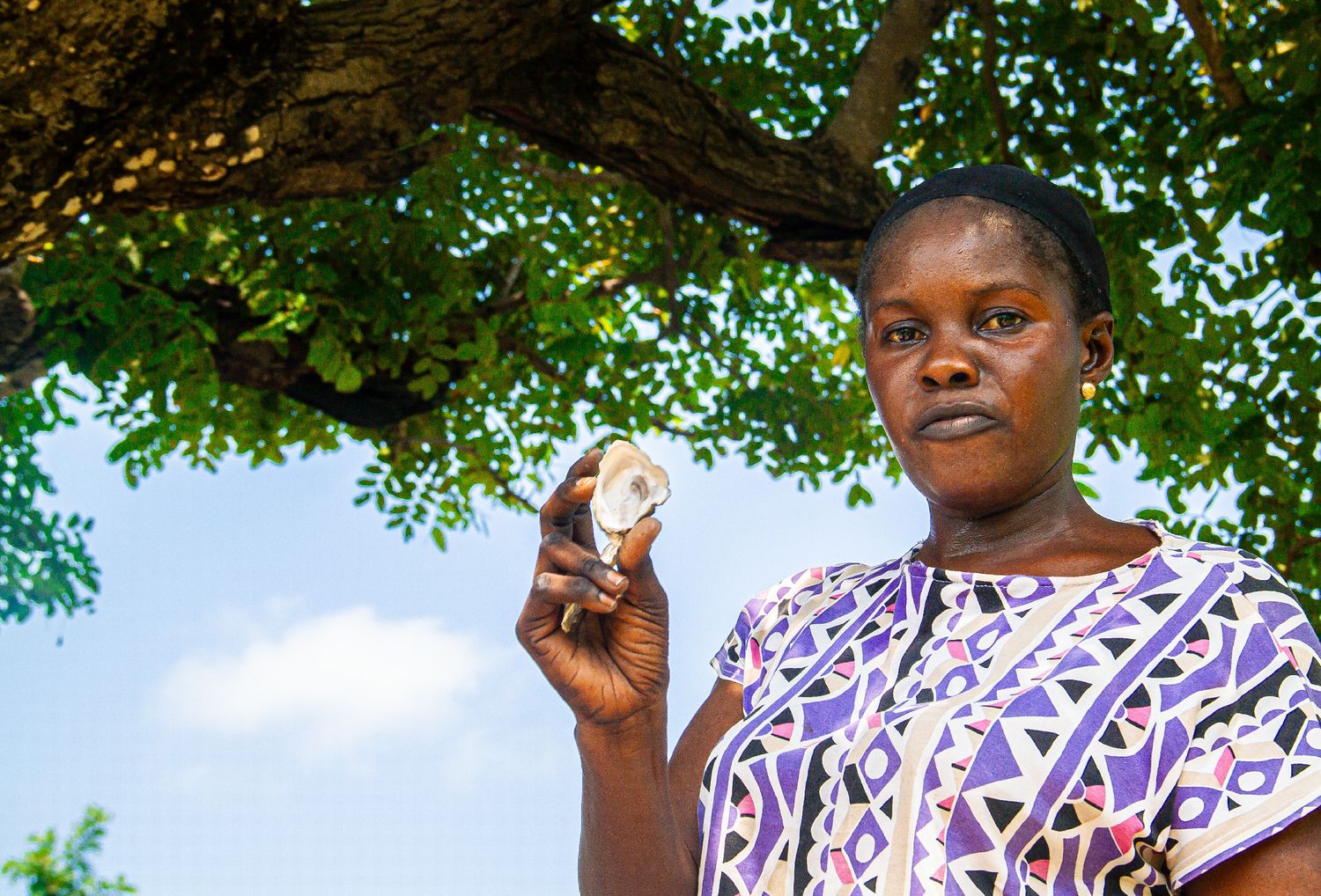 A woman holds up a small round object while standing under a large tree in an outdoor setting.