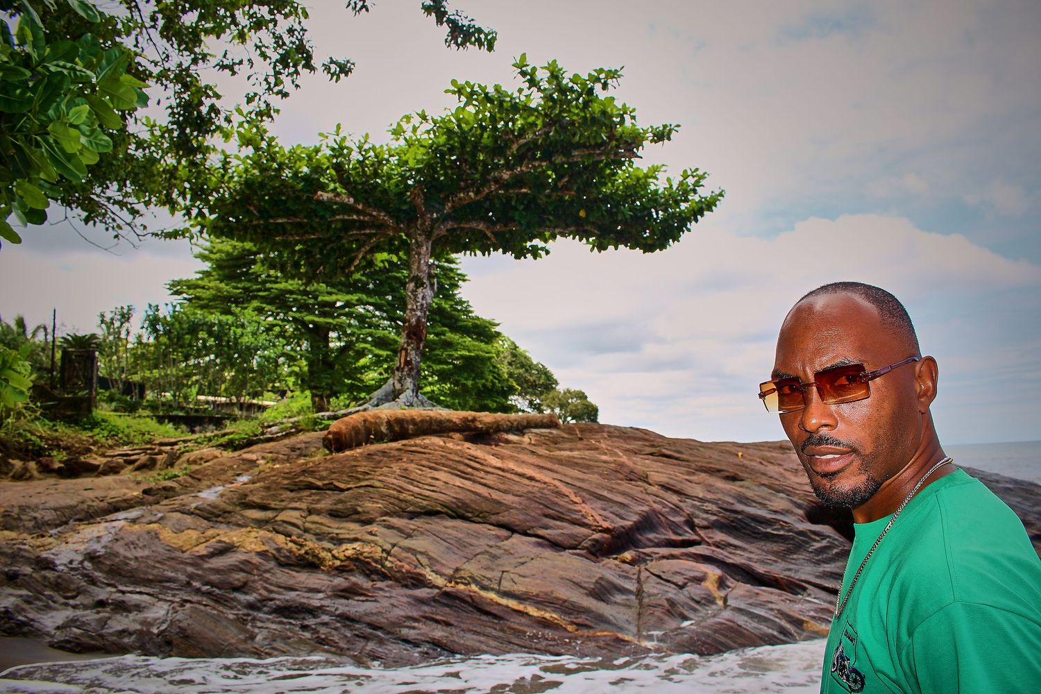 Man in green shirt posing near rocky coastal landscape with a large tree.