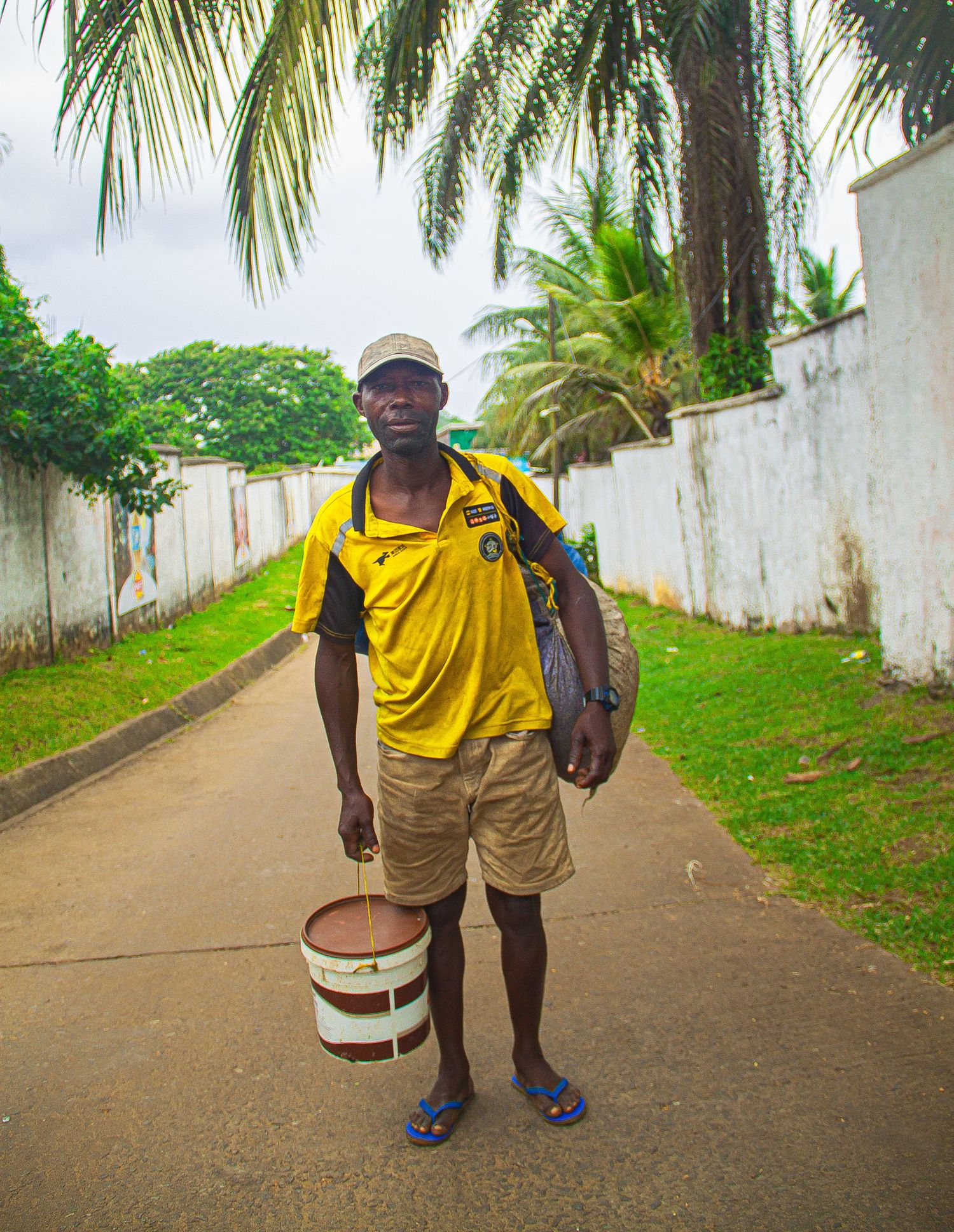 Young African man in yellow jersey holding a paint bucket while standing on a palm-lined pathway.