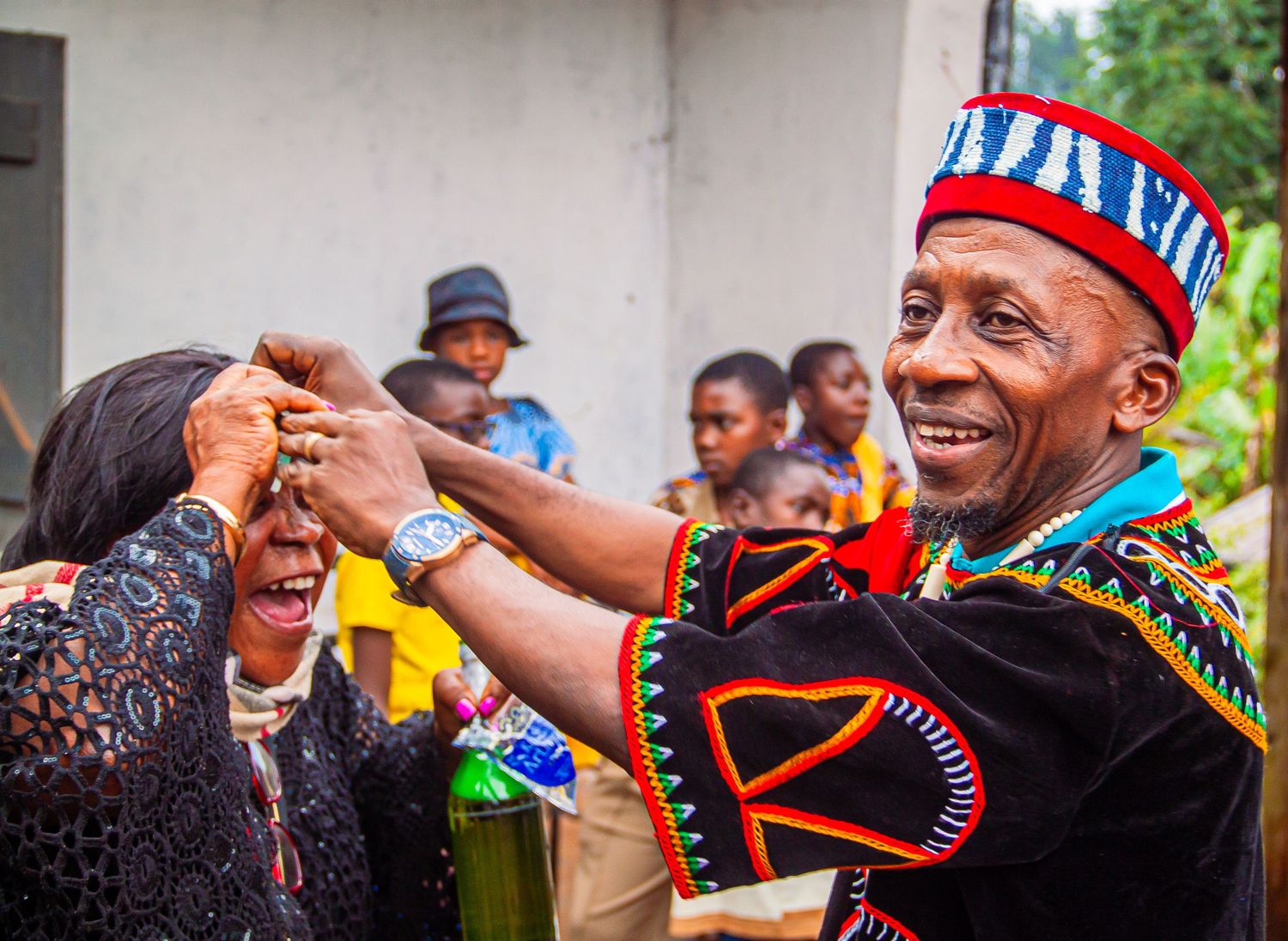 African man in traditional Cameroonian attire smiling while adjusting a woman's accessories outdoors.