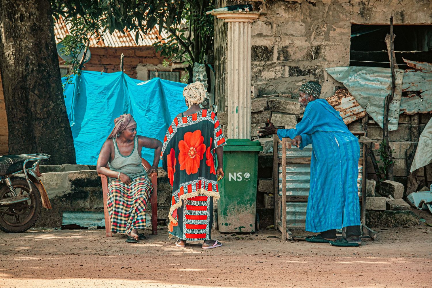 Three African women in colorful traditional dress gathered around a green waste bin outdoors.