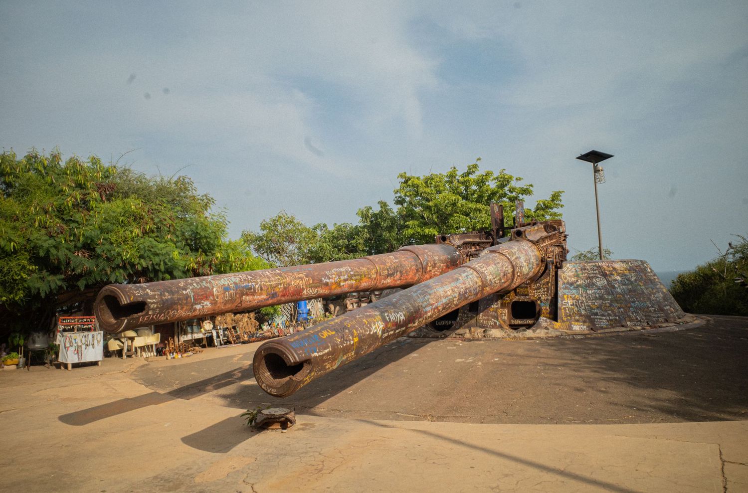 A large rusty historic cannon lies on a stone platform surrounded by trees under blue sky.