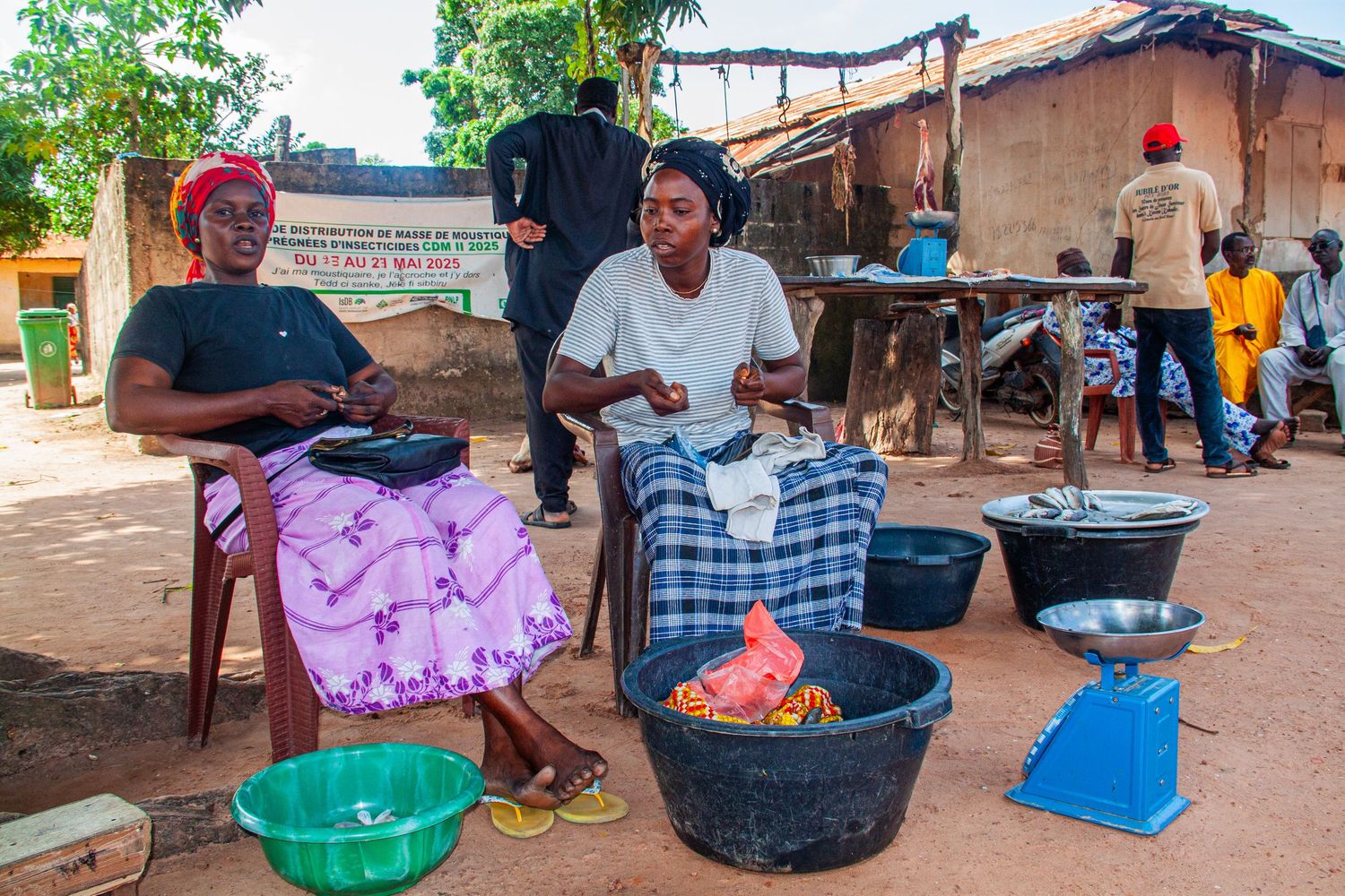Two women sell goods at an outdoor market with weighing scales in a West African village.
