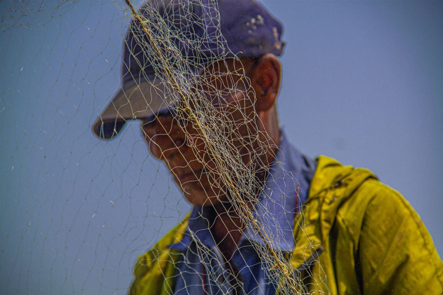 Close-up of fisherman in cap with fishing net draped over face against blue sky.