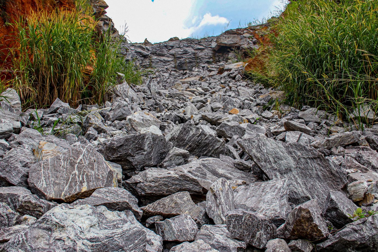 Rocky scree slope with grey boulders flanked by green and orange grass under a blue sky.