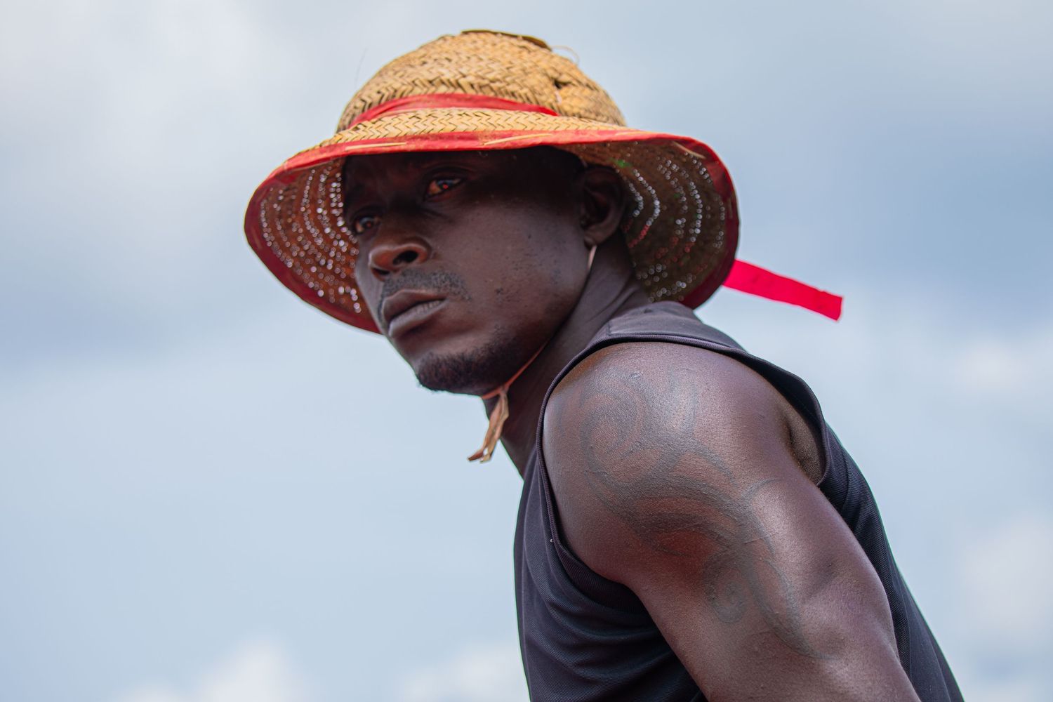 A young man wearing a straw hat looks up against a cloudy sky outdoors.