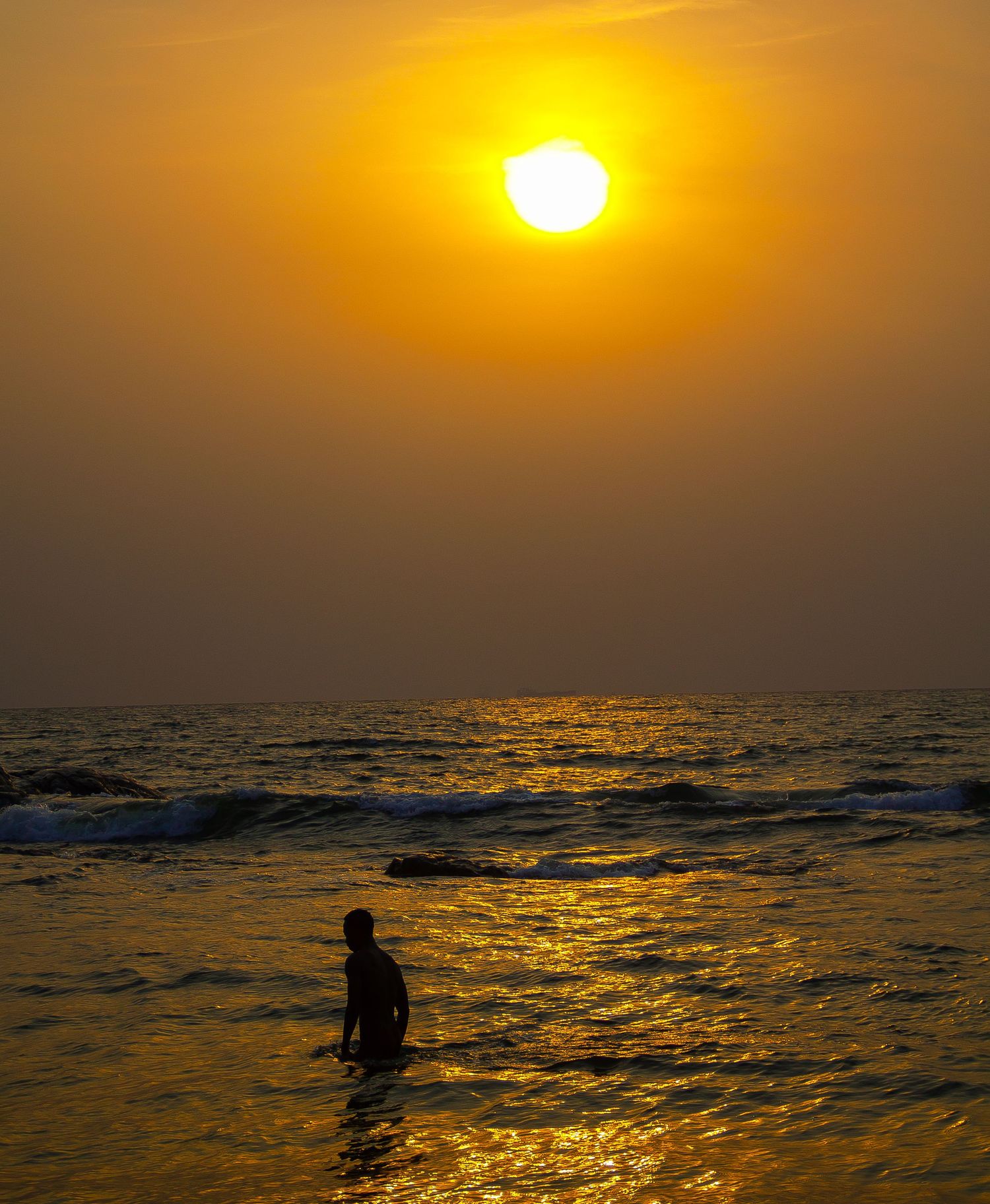 Lone silhouette of a person wading in ocean waves beneath a vibrant golden sunset sky.