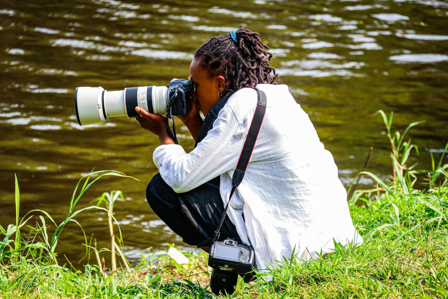 A woman crouches by a riverbank, capturing photos with a professional camera fitted with a large telephoto lens.