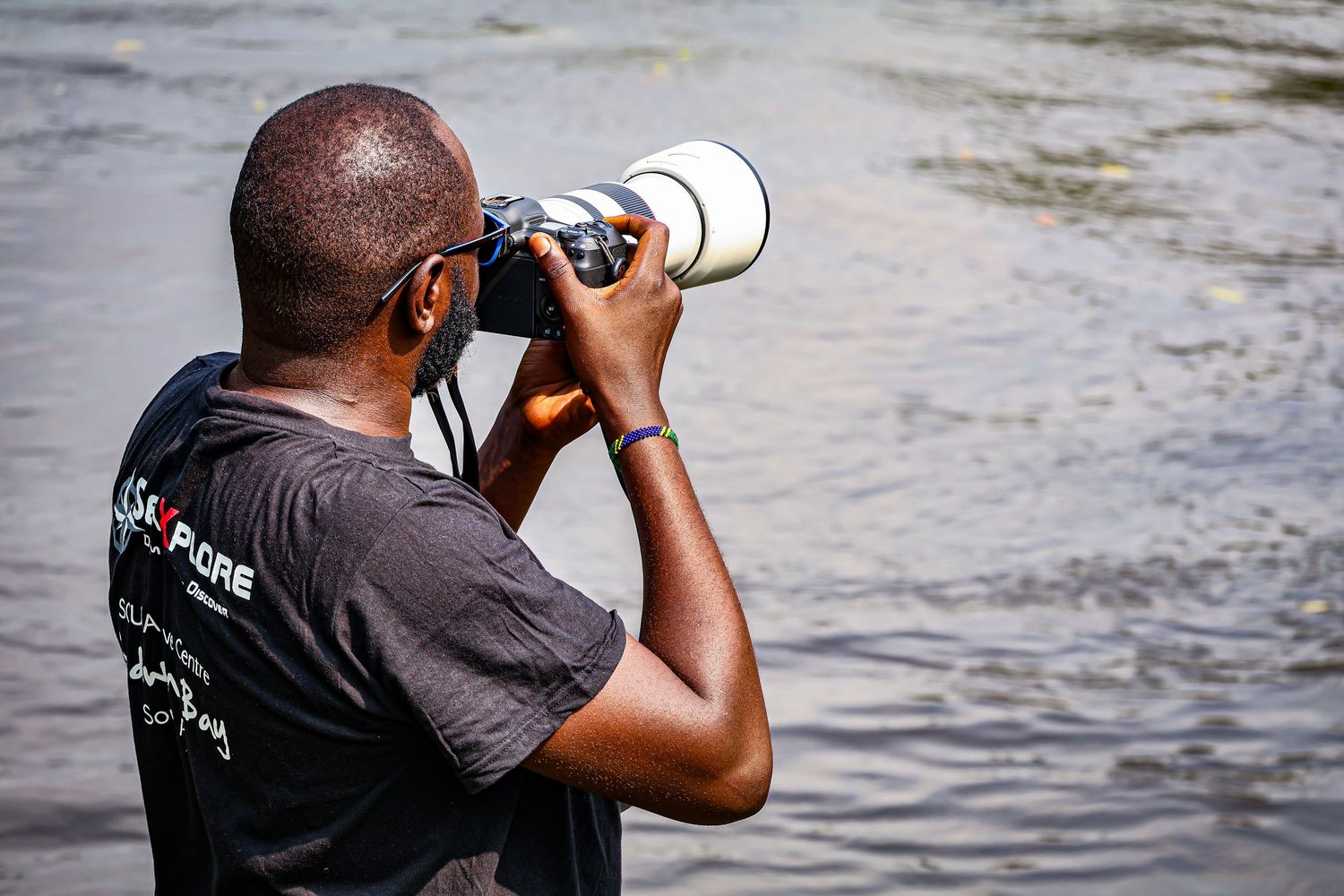 A man photographs wildlife near a river using a professional camera with a large white telephoto lens.