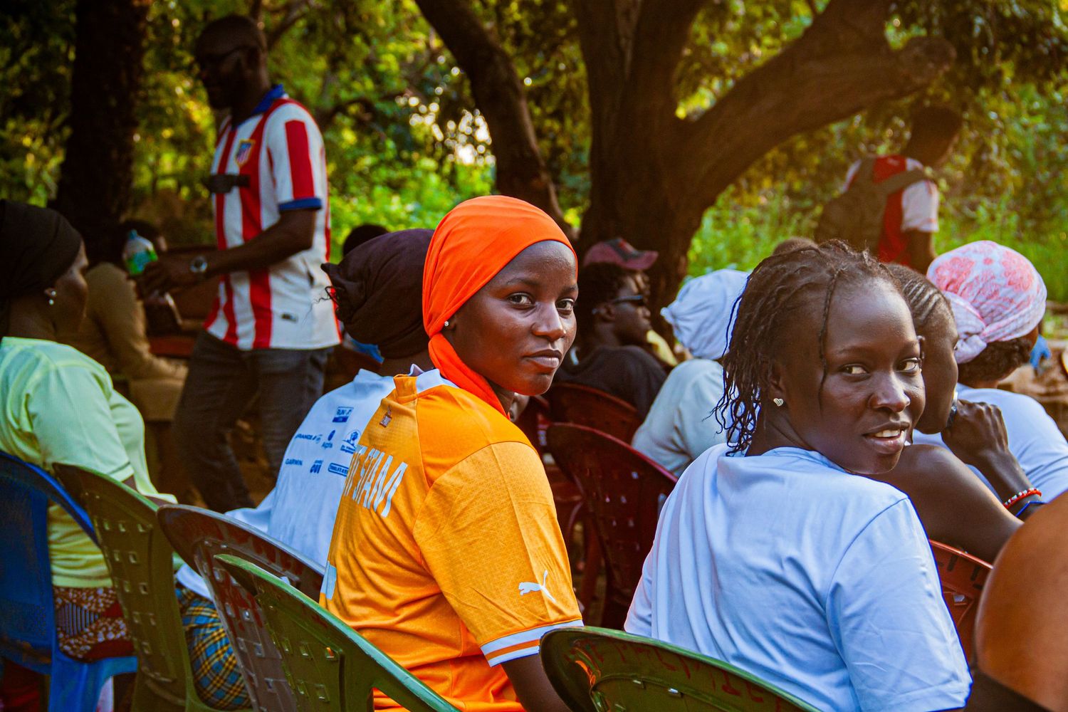 Young African girl in orange hijab and jersey seated outdoors at a community gathering.