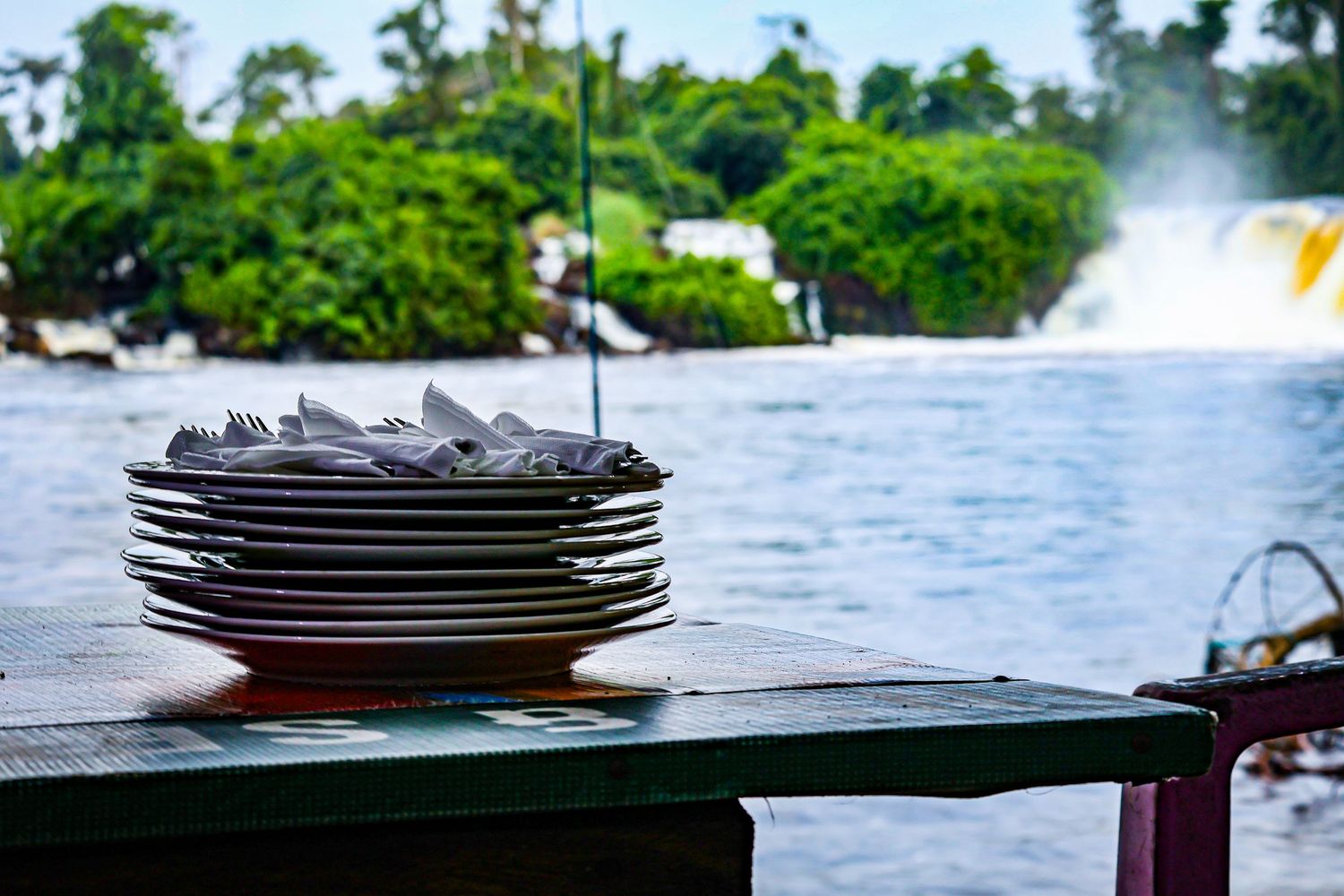 Stack of white plates and cutlery on a riverside restaurant table with tropical waterfall in background.
