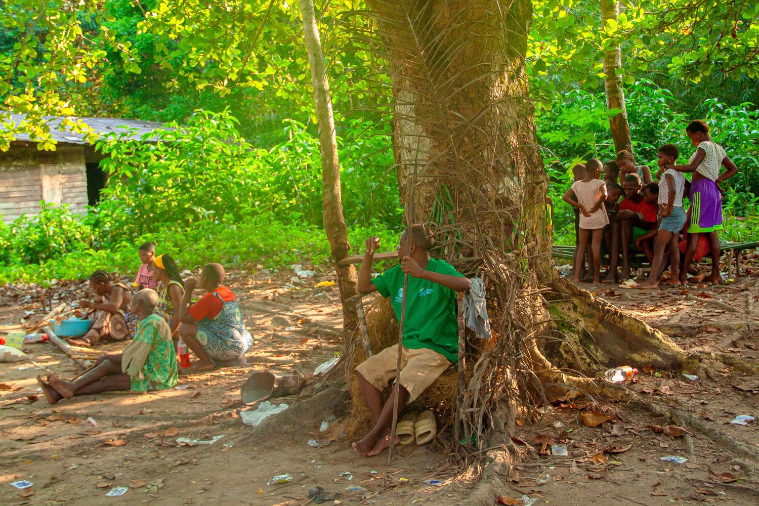 Children and adults gathering around a large tree in a tropical African village setting.