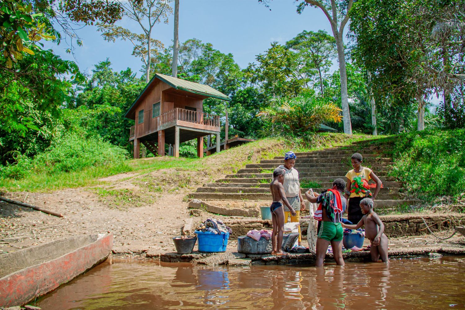 Locals doing laundry at river's edge with elevated wooden house on stilts and stone steps in background.