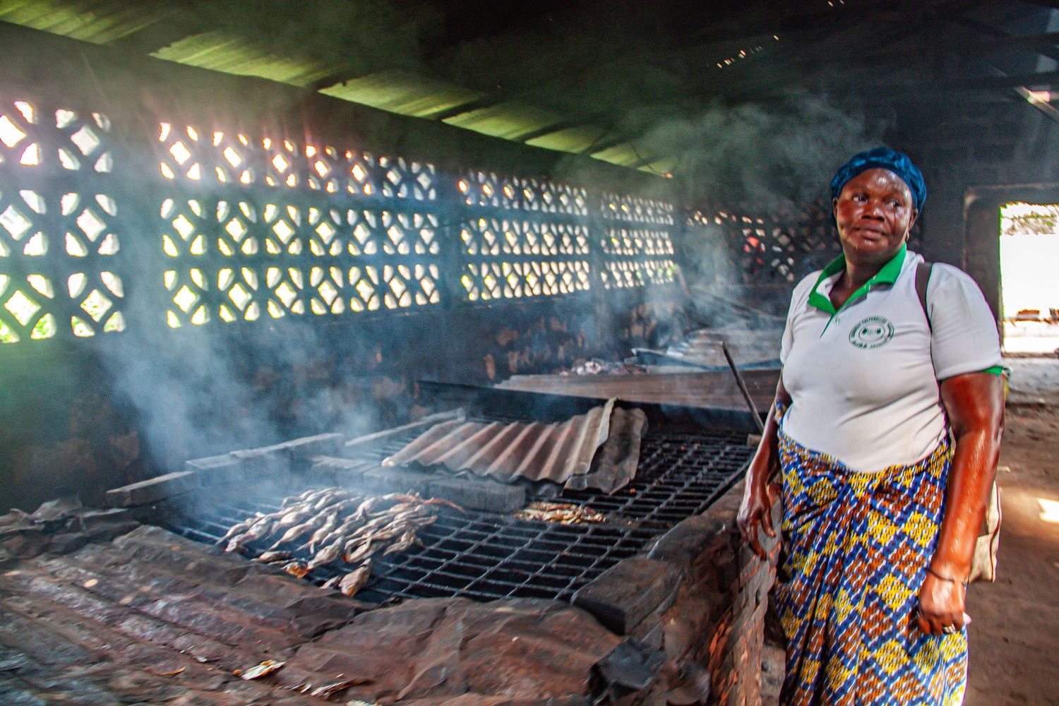 A woman stands beside a large smoking fish grill in a traditional African smokehouse.