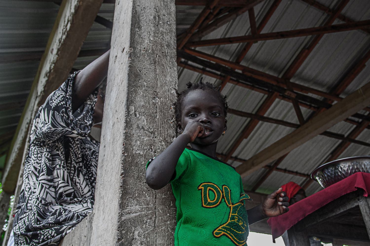 Young African toddler in green shirt standing by wooden post under corrugated roof.