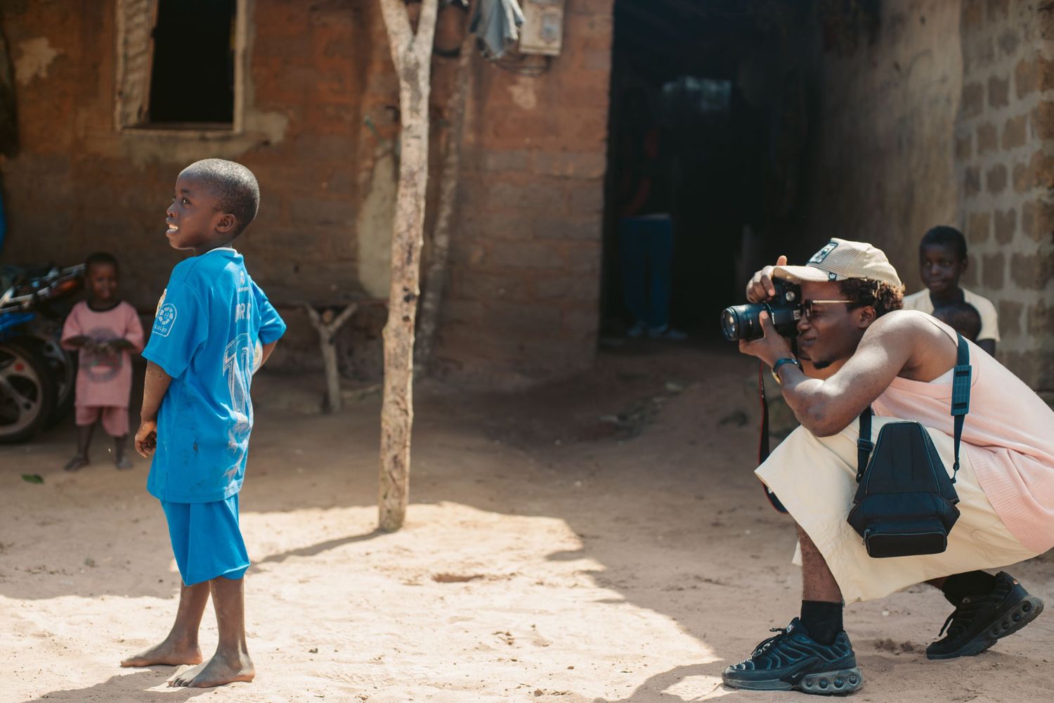 A photographer crouches to capture a portrait of a young child in a rural African village setting.