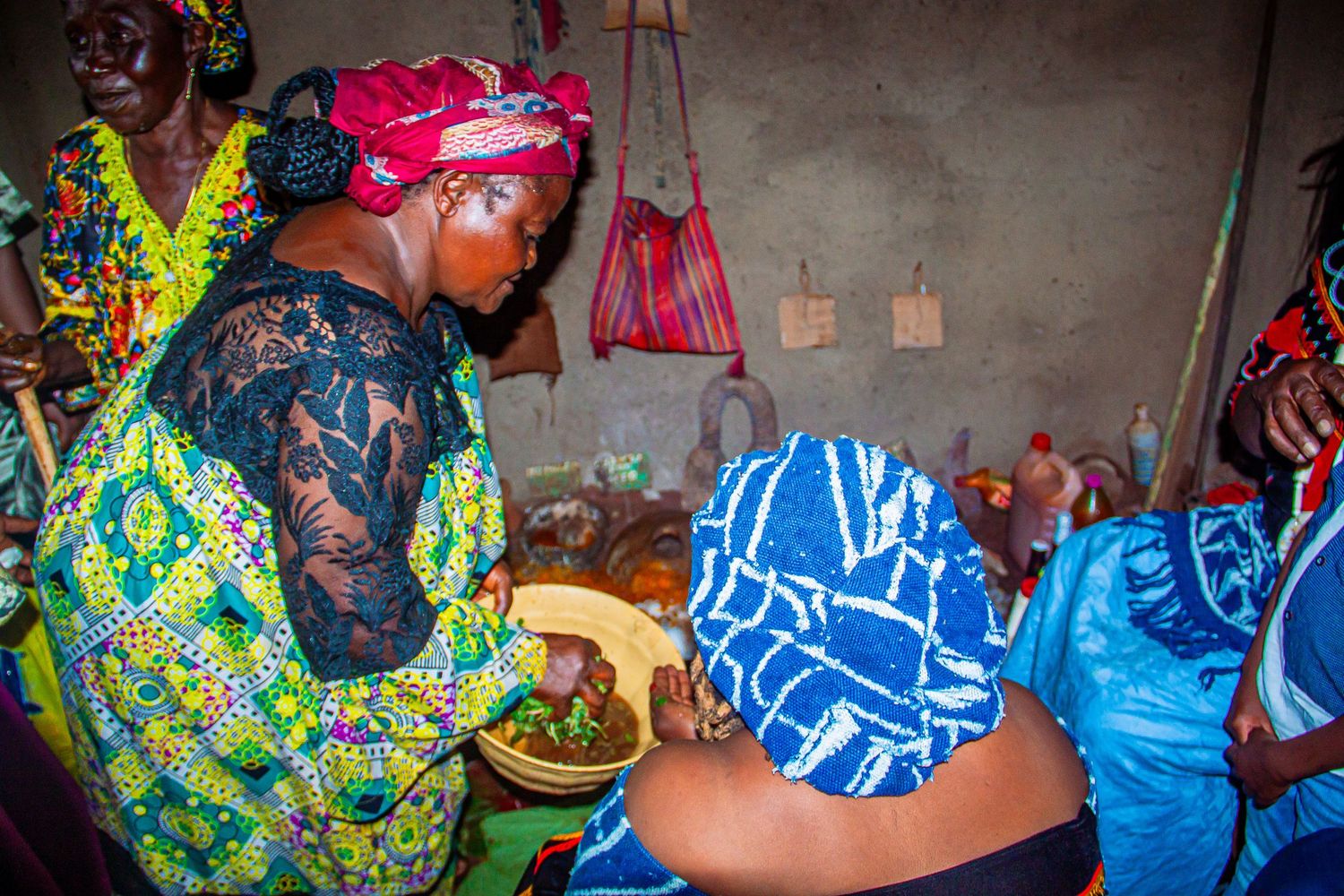 Women in vibrant African attire gather around food at a traditional cultural celebration indoors.