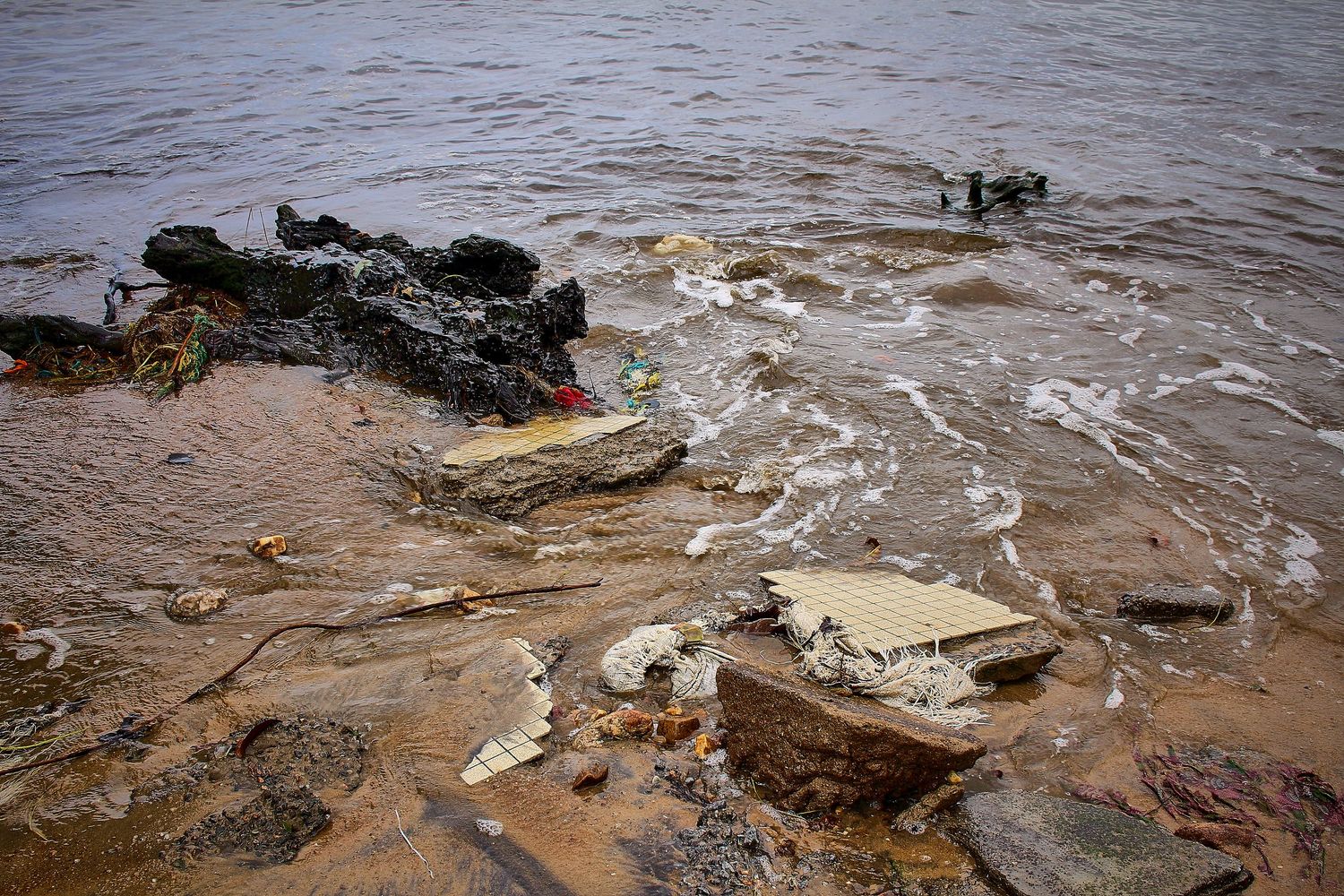 Debris and old tires scattered along a rocky shoreline with murky water.