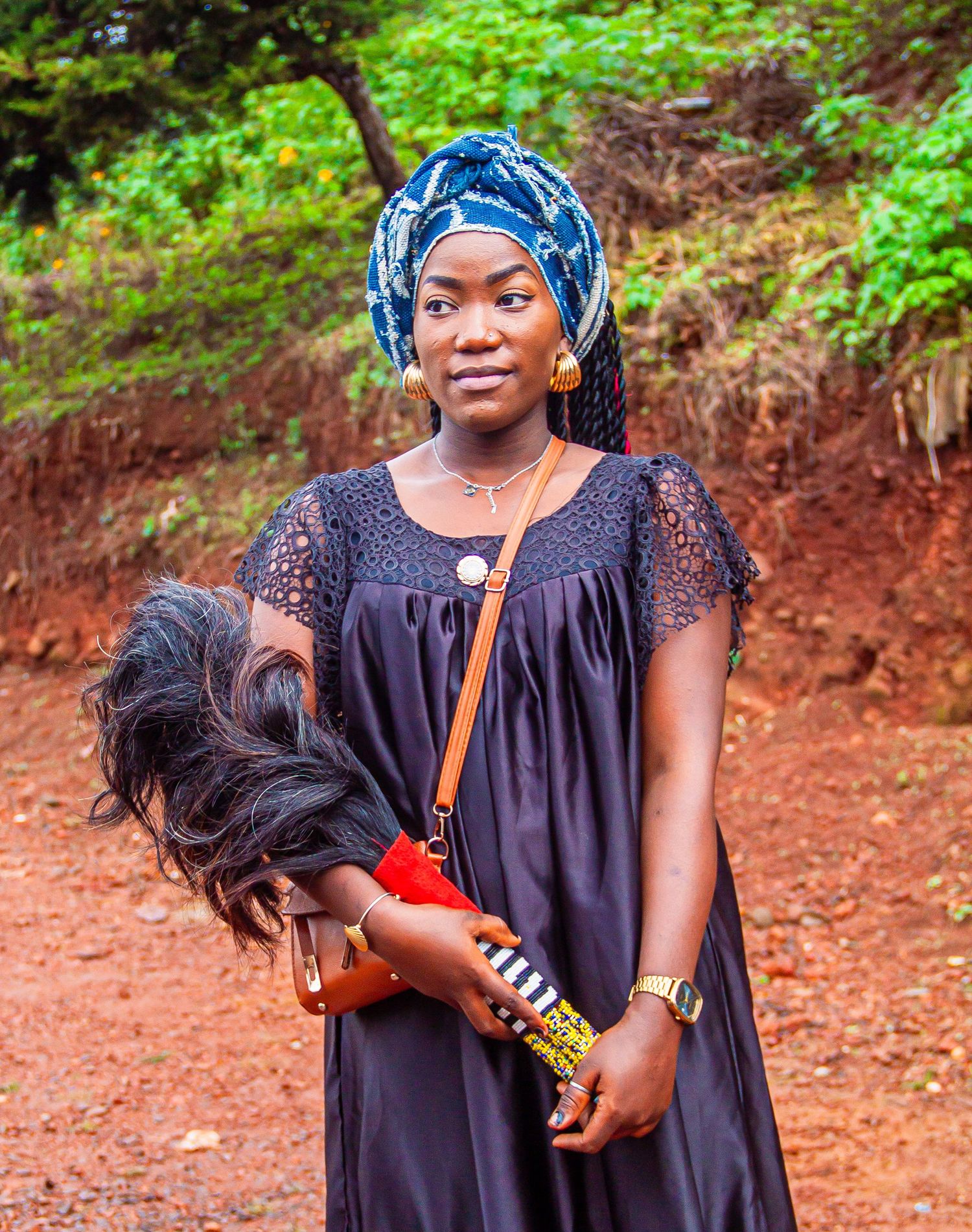 Young African woman in blue head wrap and black dress holds hair piece outdoors on red dirt path.