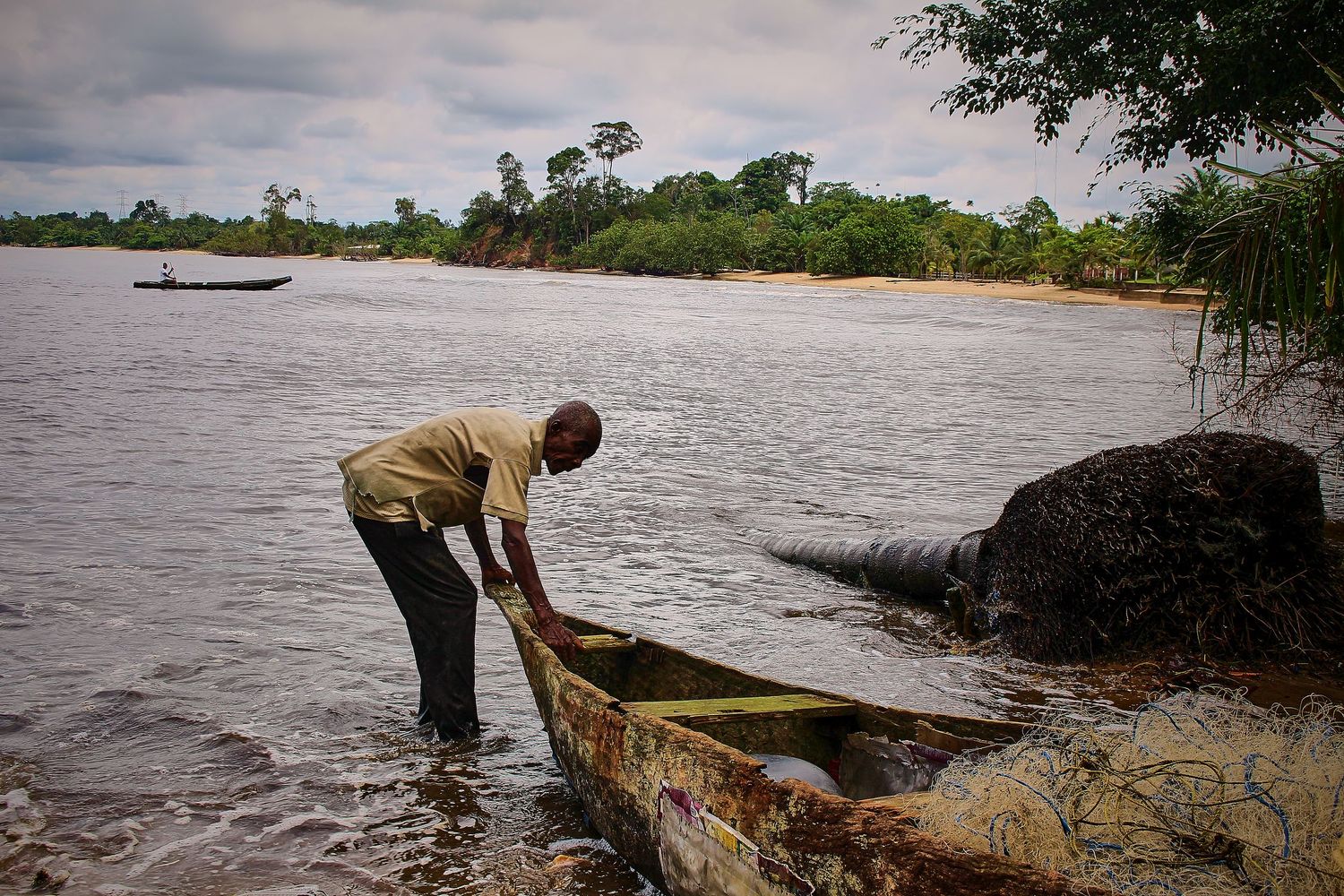 A man tends to a weathered wooden canoe at the edge of a tropical river surrounded by lush greenery.