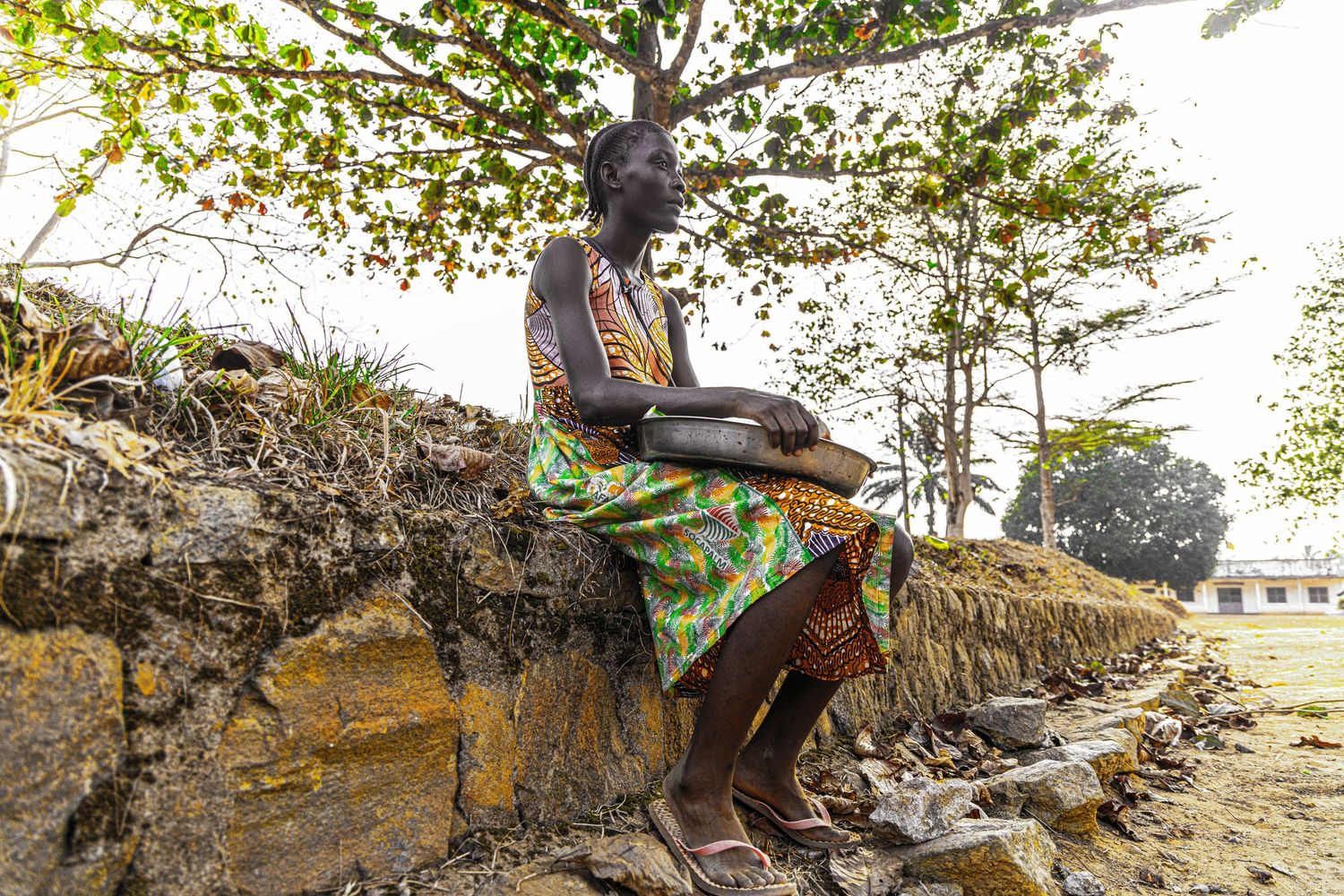 African woman in colorful dress sitting on a stone wall under a tree.