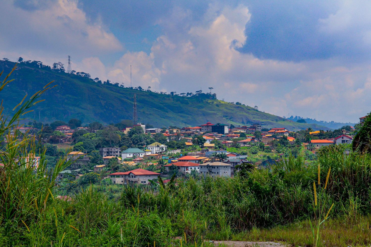 Lush green hillside town with colorful houses nestled among tropical vegetation under a cloudy sky.