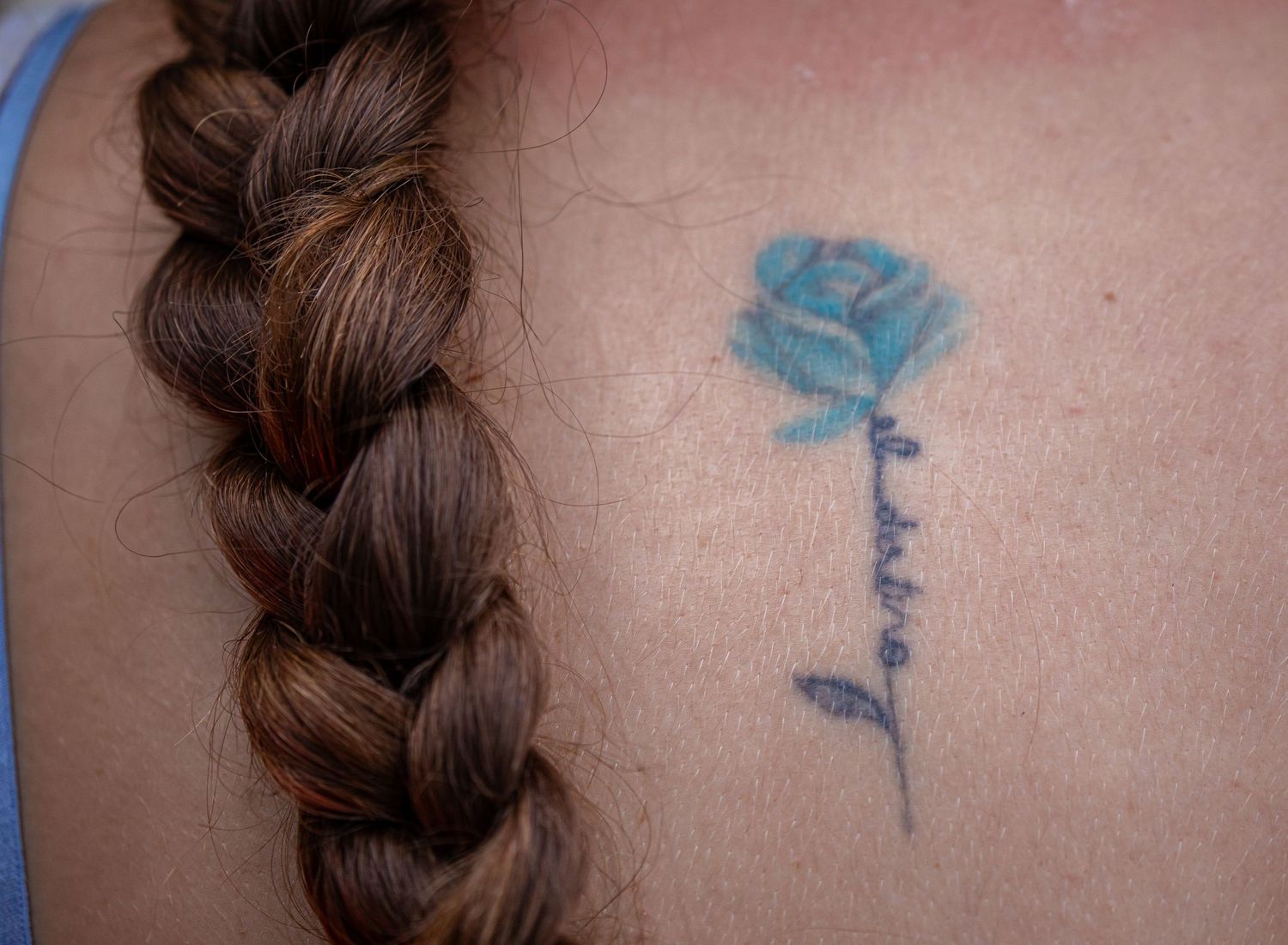 Blue rose tattoo with script lettering on a person's back next to a braided hairstyle.