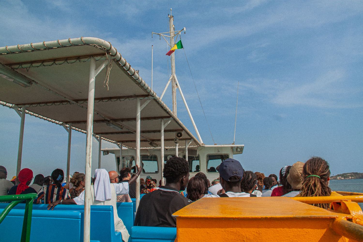 Tourists on a ferry boat deck with Senegalese flag flying against a blue sky over calm ocean waters.