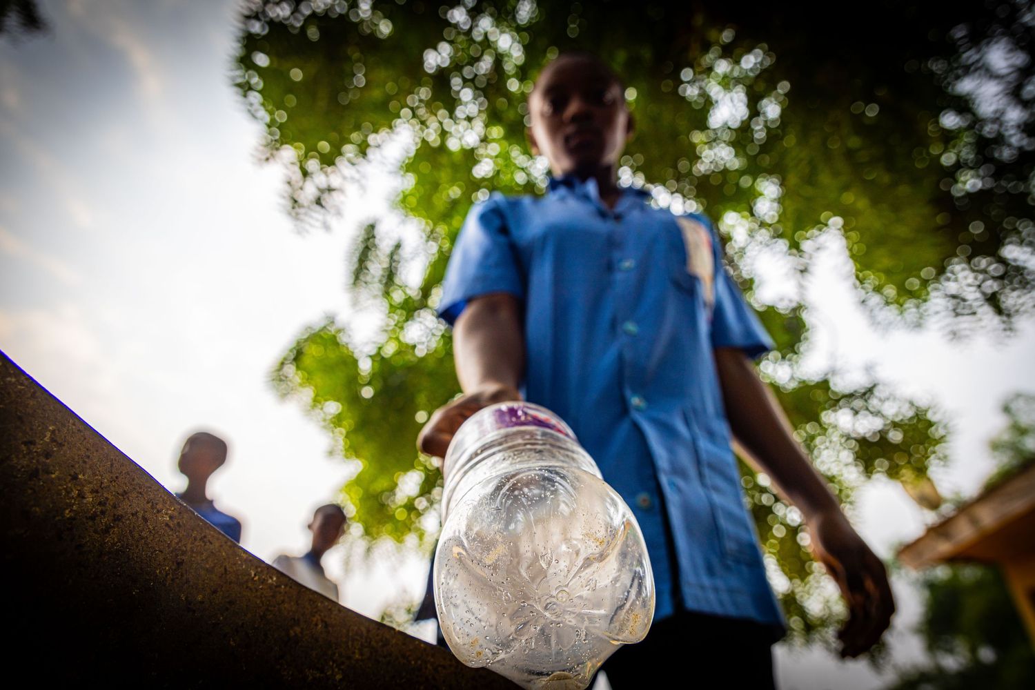 Child in blue uniform holding a plastic bottle outdoors under green trees.