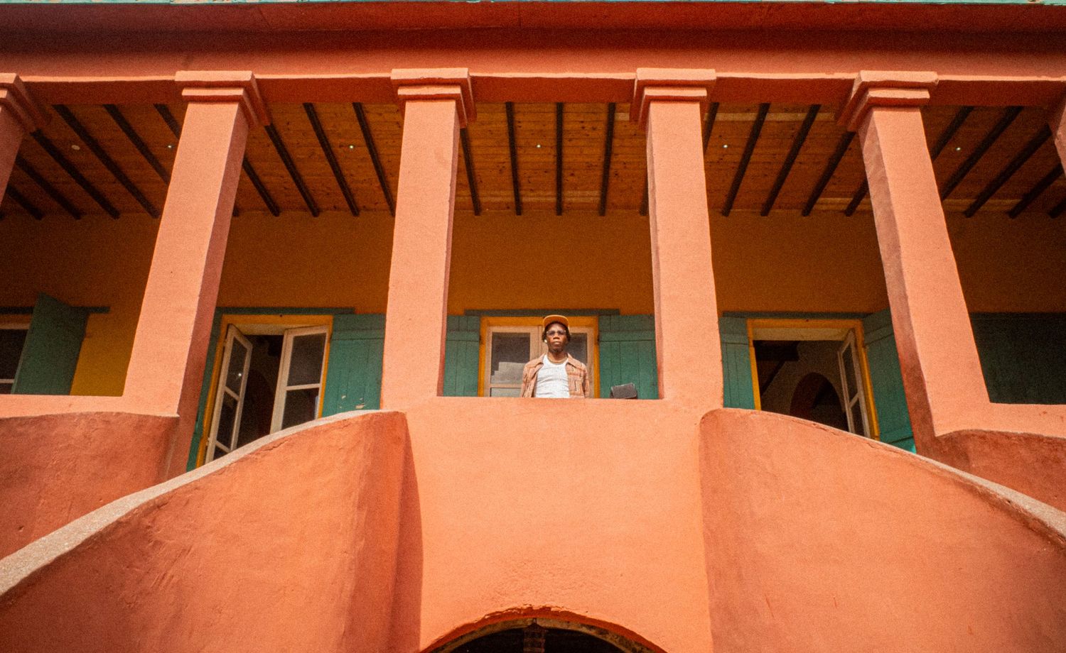 A person stands on the balcony of a terracotta-colored colonial building with green shuttered windows.