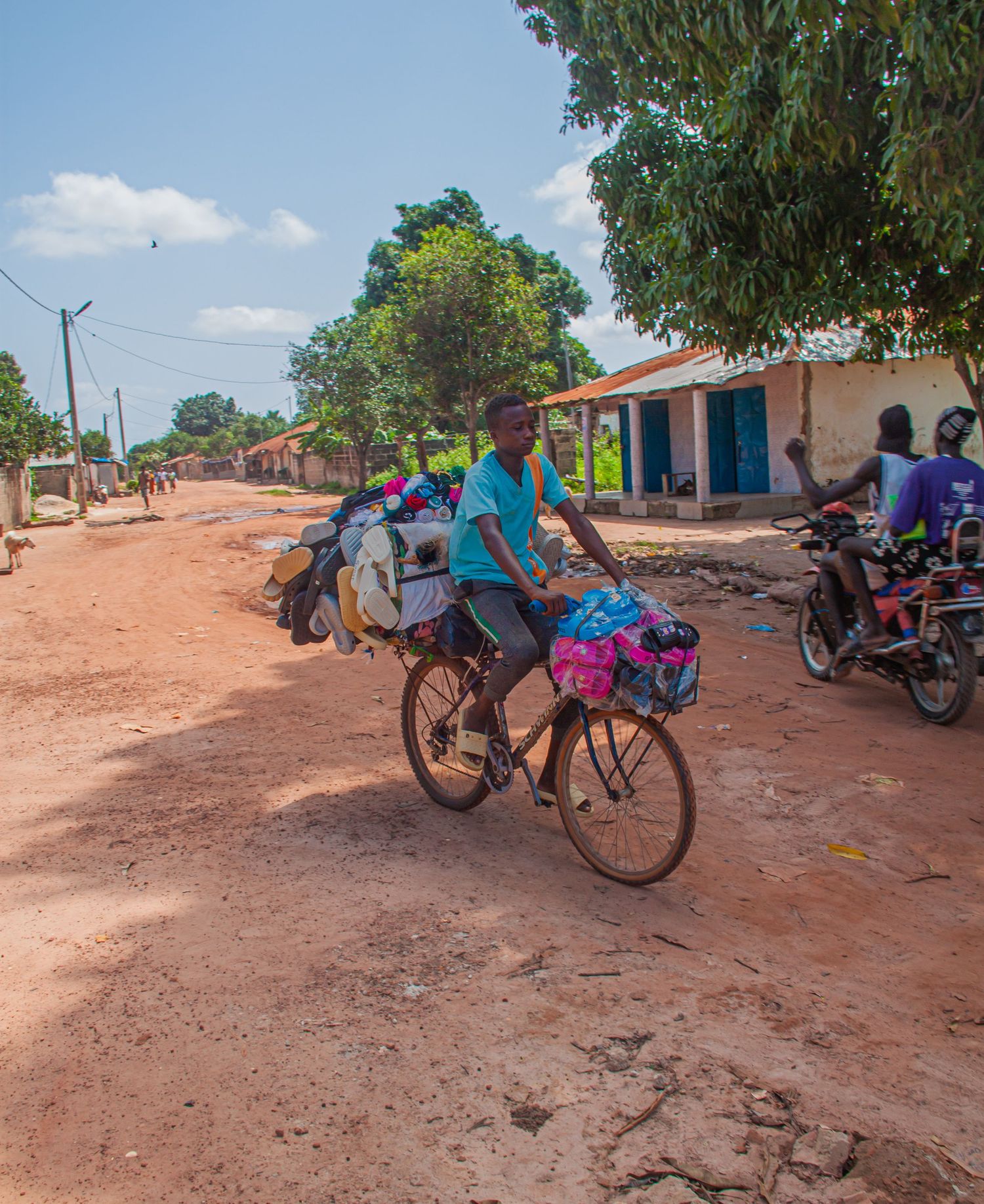 Man cycling along a dusty red dirt road carrying heavy loads of goods in a rural African village.