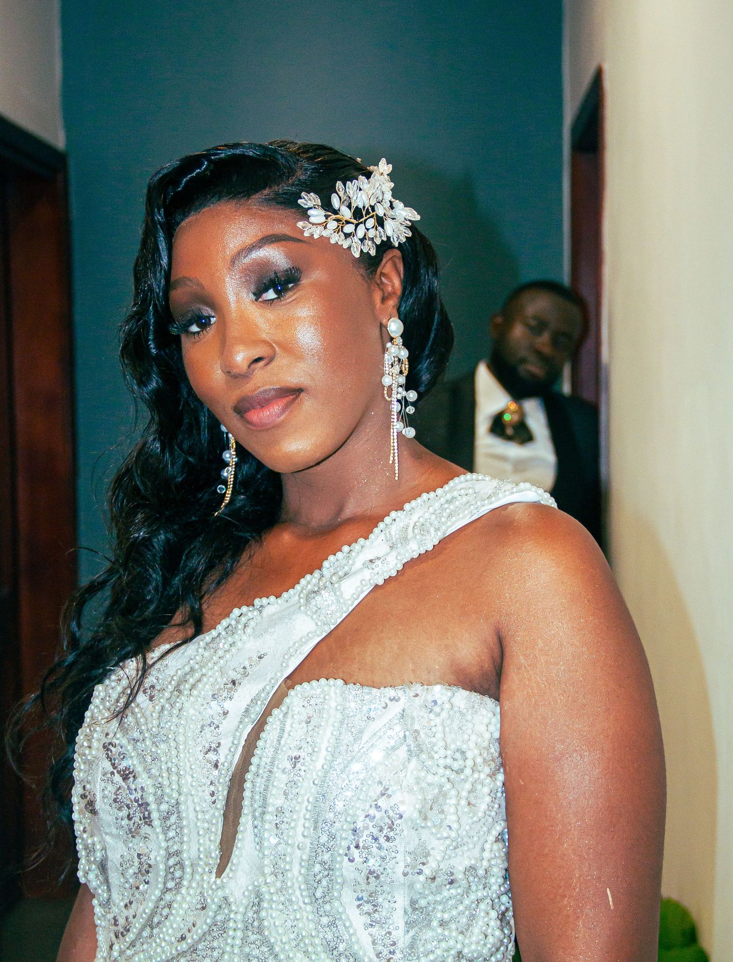 Bride in beaded white gown with floral hair accessory and chandelier earrings posing indoors.