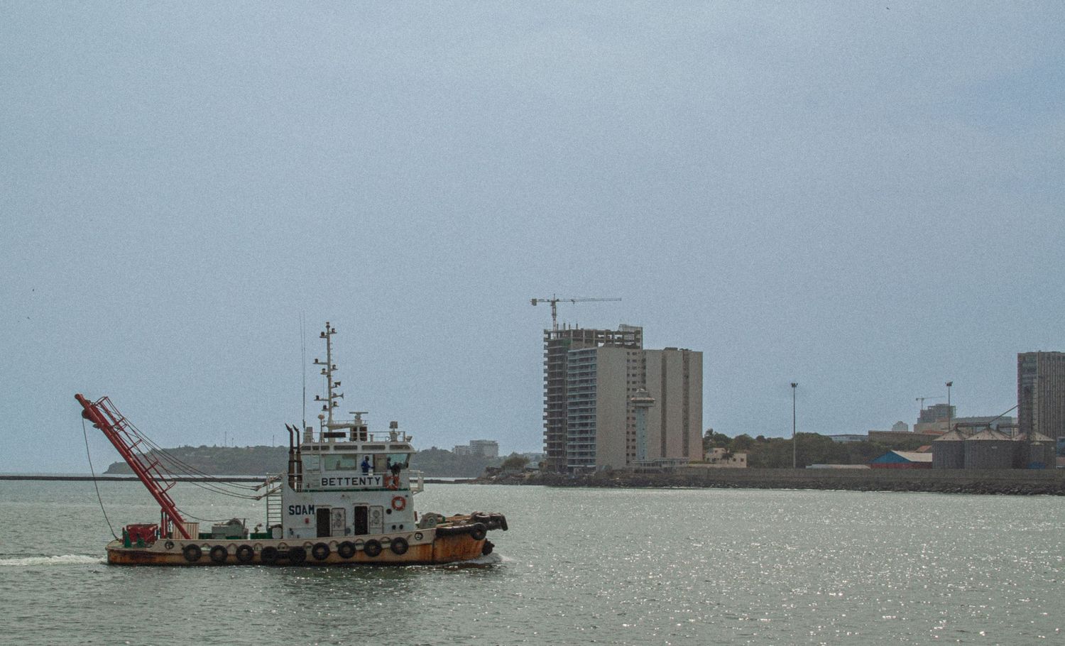 Industrial tugboat with crane sailing past urban waterfront with tall buildings under overcast sky.