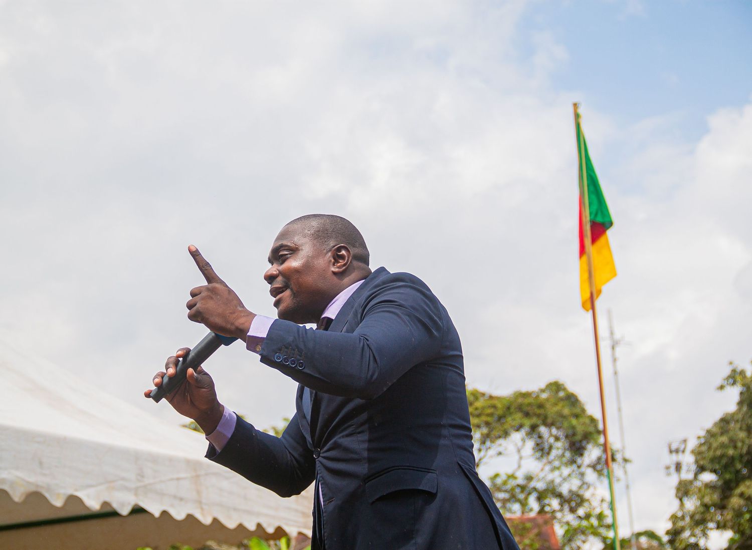 African man in blue suit speaking into microphone at outdoor event with colorful flag in background.