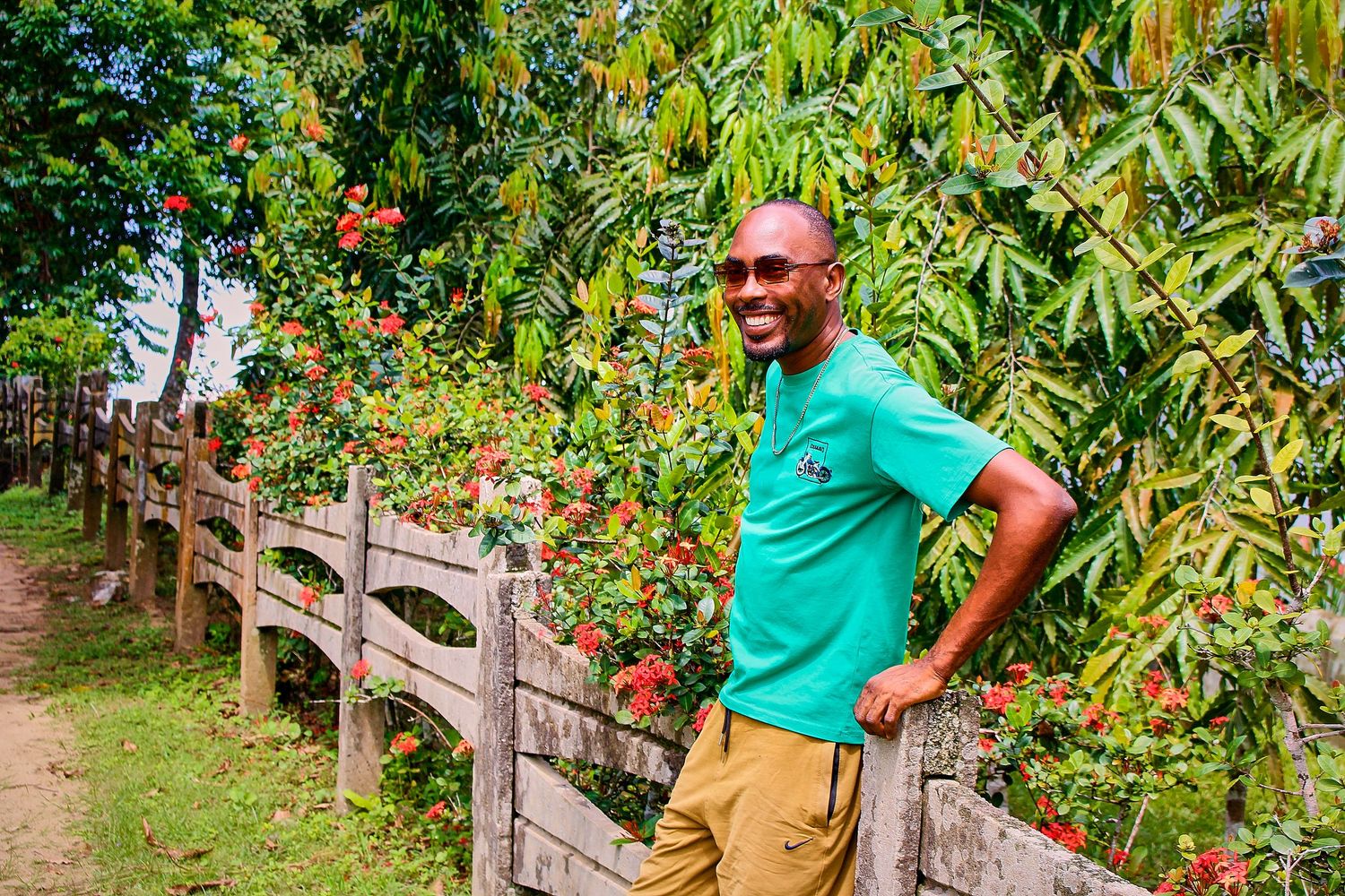 A smiling man in a green shirt poses beside a wooden fence adorned with vibrant tropical flowers.