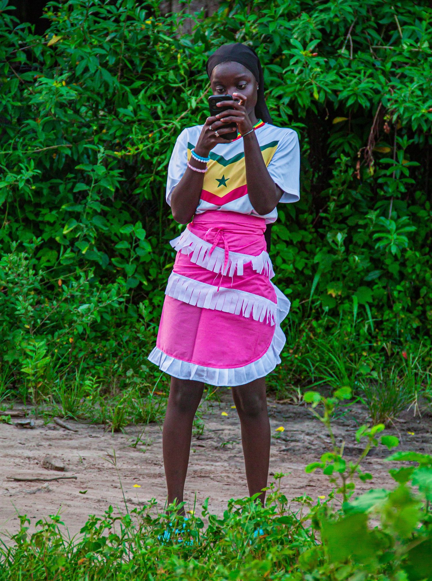 Young African girl in pink fringe skirt and Ghana flag shirt standing in lush green surroundings.