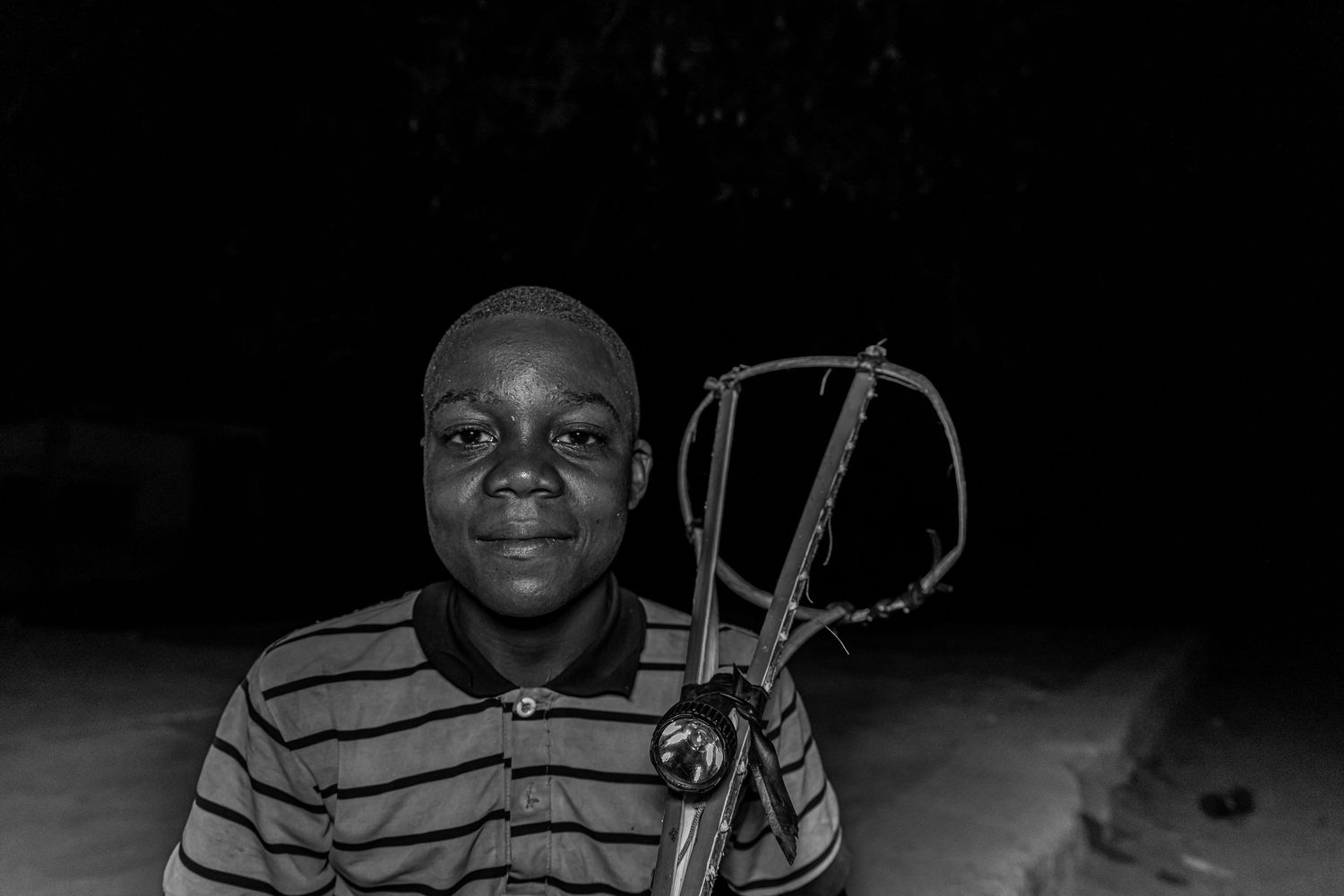 Black and white portrait of a young African boy holding a handmade toy car at night.