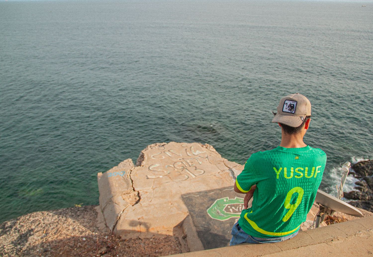 Person in green Yusuf jersey sitting on rocky cliff edge overlooking calm blue ocean waters.