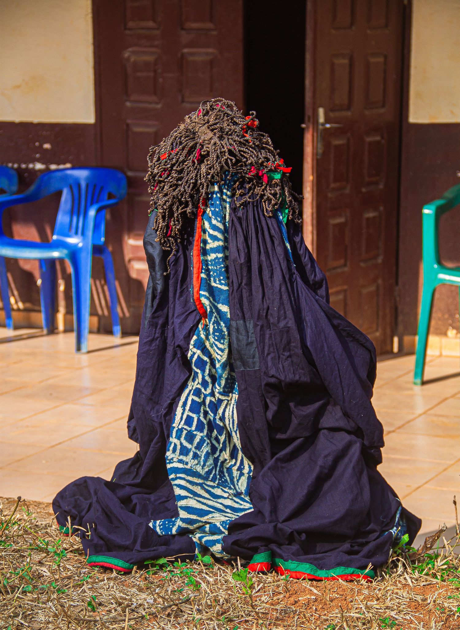 Person in traditional African ceremonial costume with dark blue robes and decorative headdress sitting on ground.