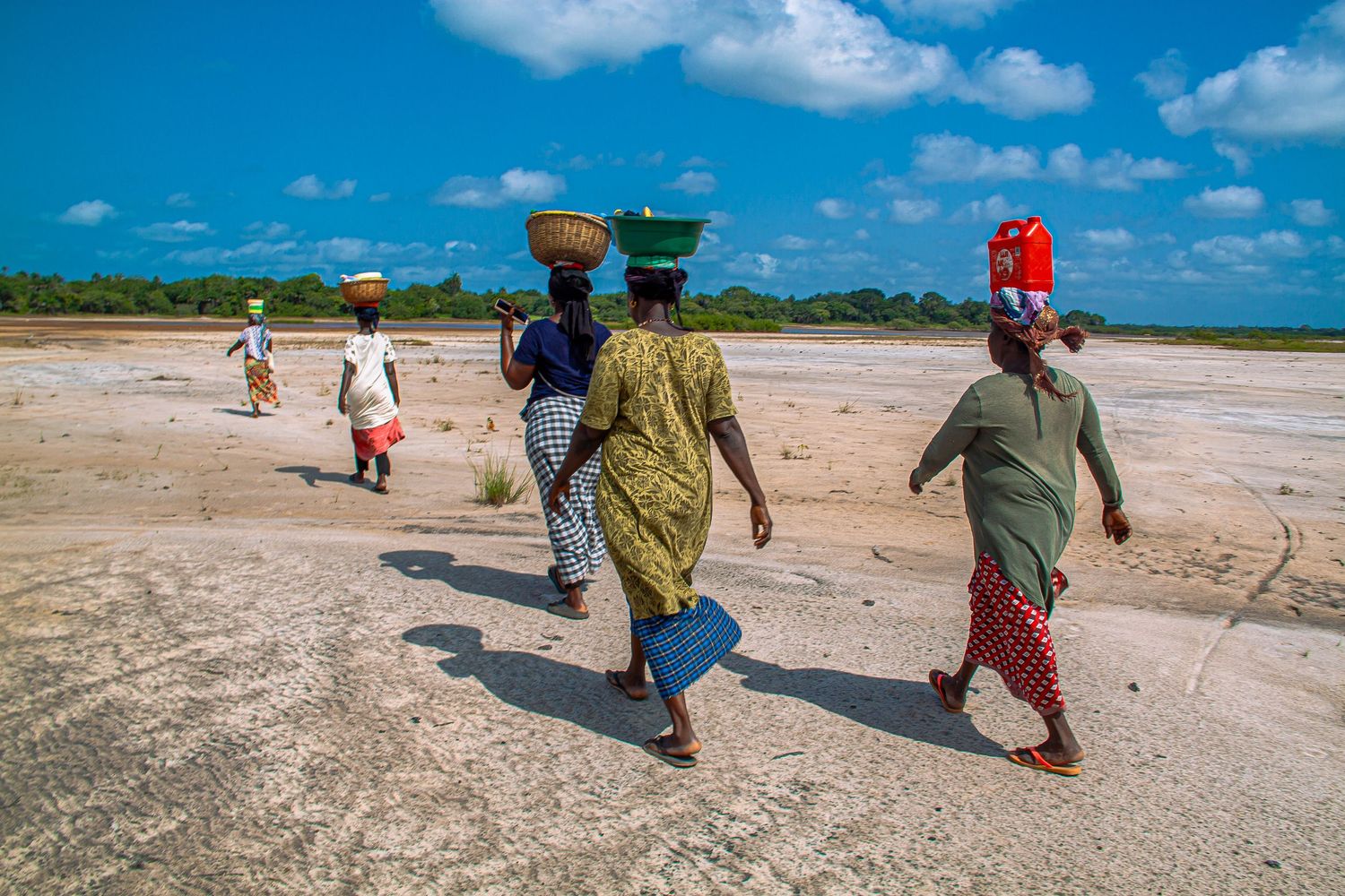African women carry baskets and containers on their heads across a dry sandy landscape under blue sky.
