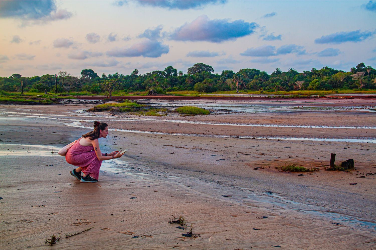 Woman crouching on sandy tidal flats at sunset near a lush African riverbank.