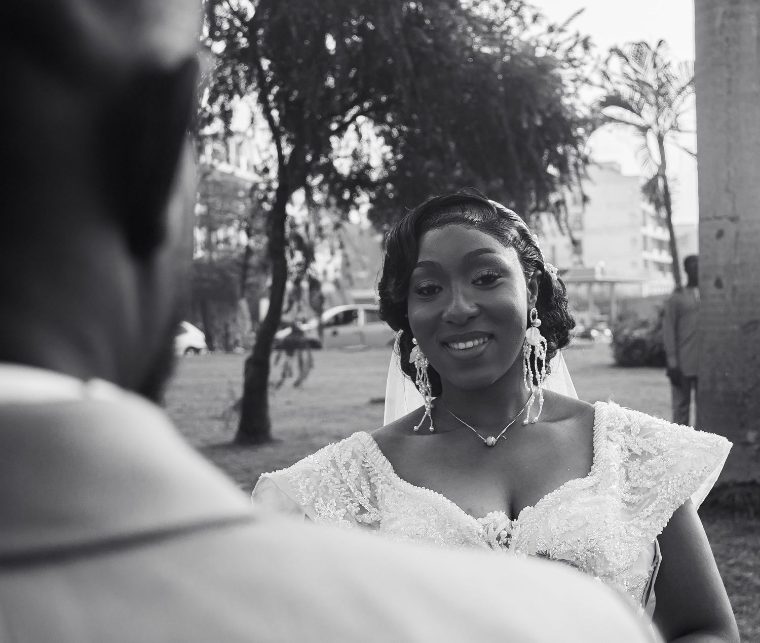Joyful African bride in white lace dress smiling at groom outdoors in black and white photo.