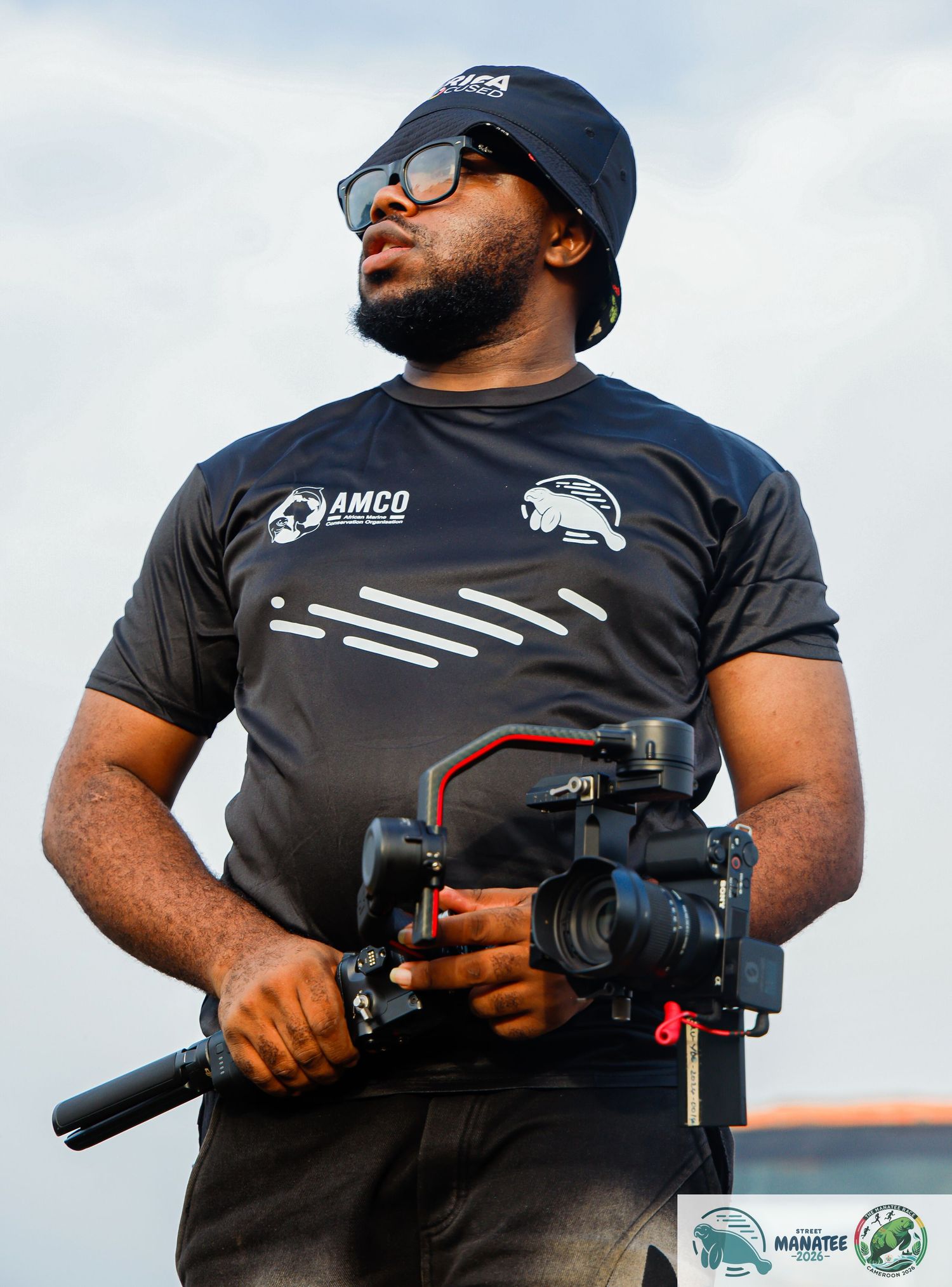 A bearded man in an AMCO black shirt holds a professional camera gimbal outdoors.