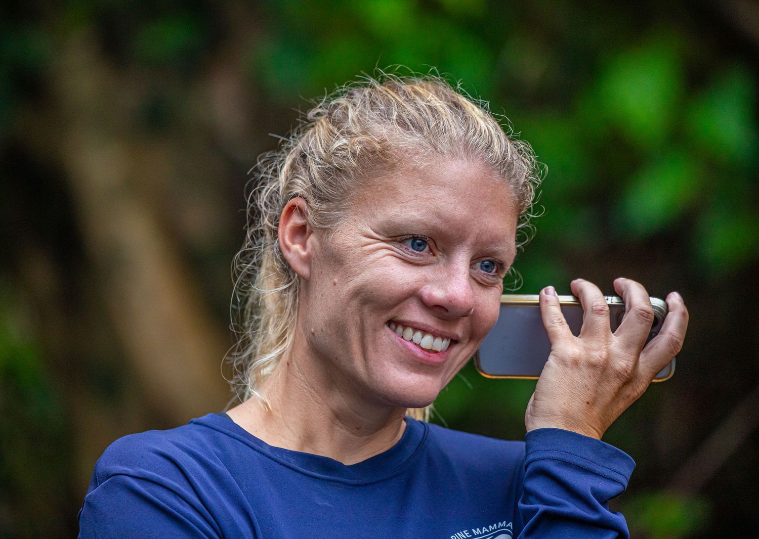Smiling blonde woman holding a smartphone to her face outdoors in a lush green setting.