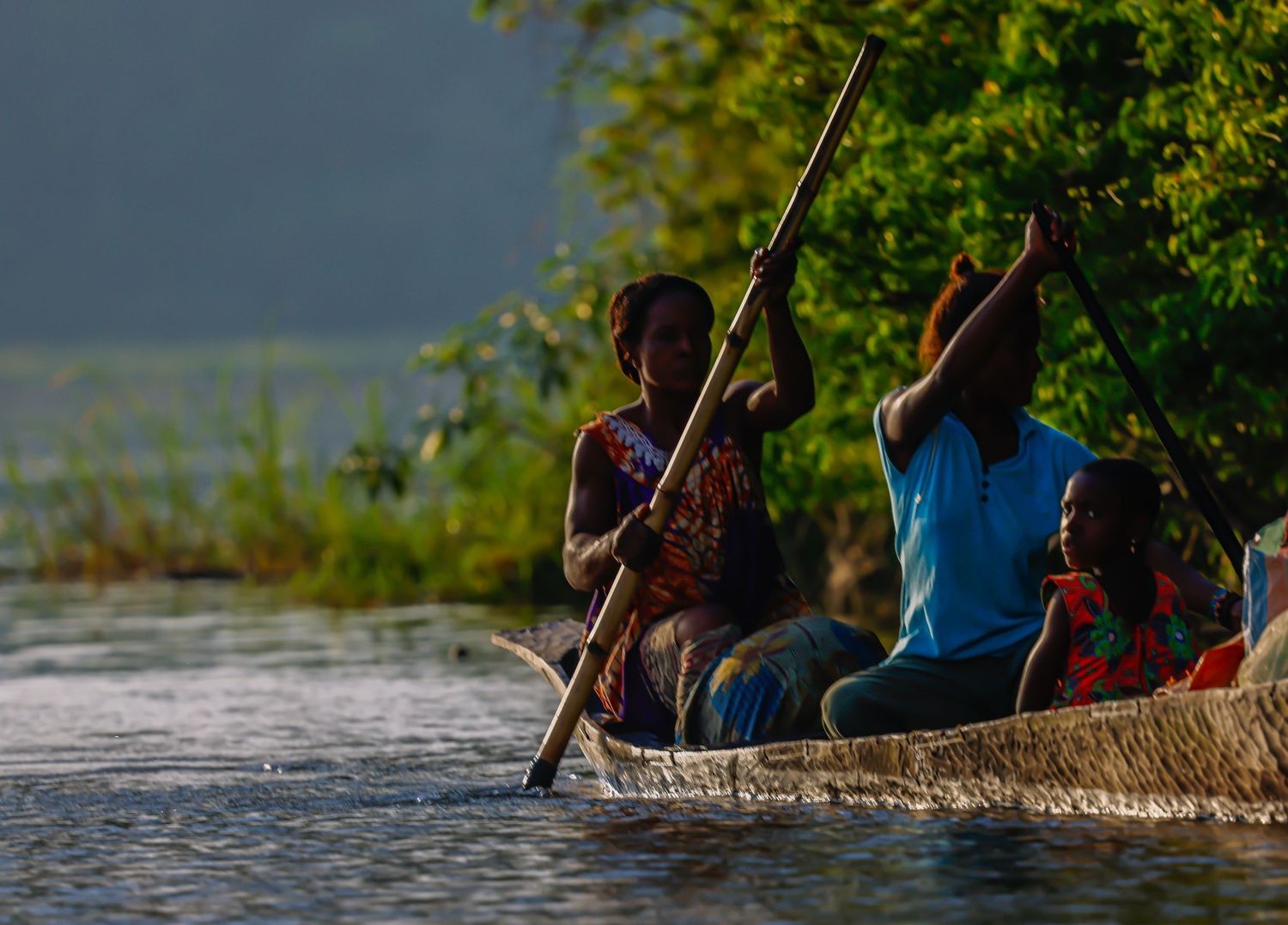 African family navigating a traditional reed boat on a misty river at golden hour with a paddle.